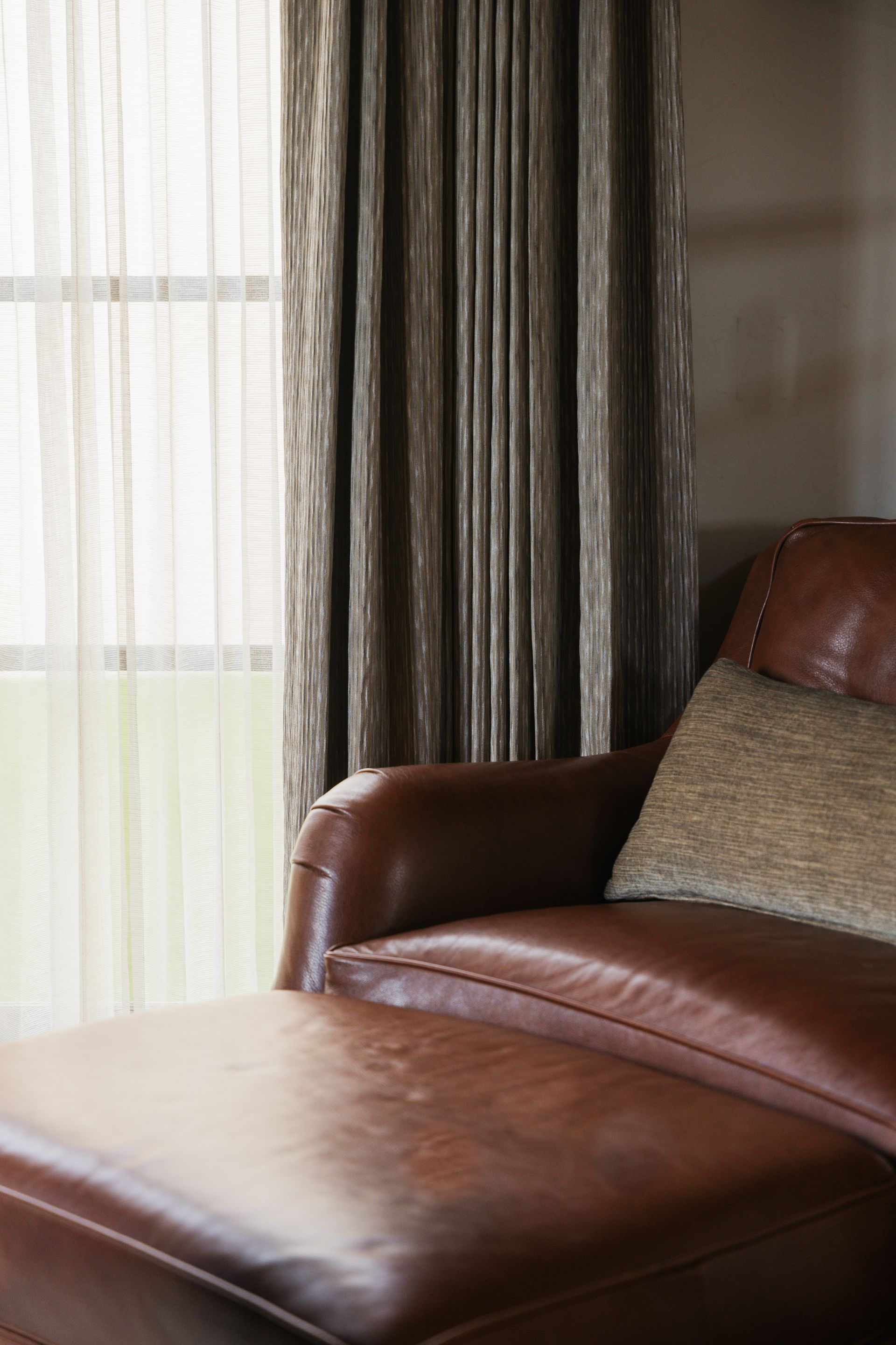 Leather chaise lounge with a decorative pillow, next to a window with sheer curtains and draped brown curtains.