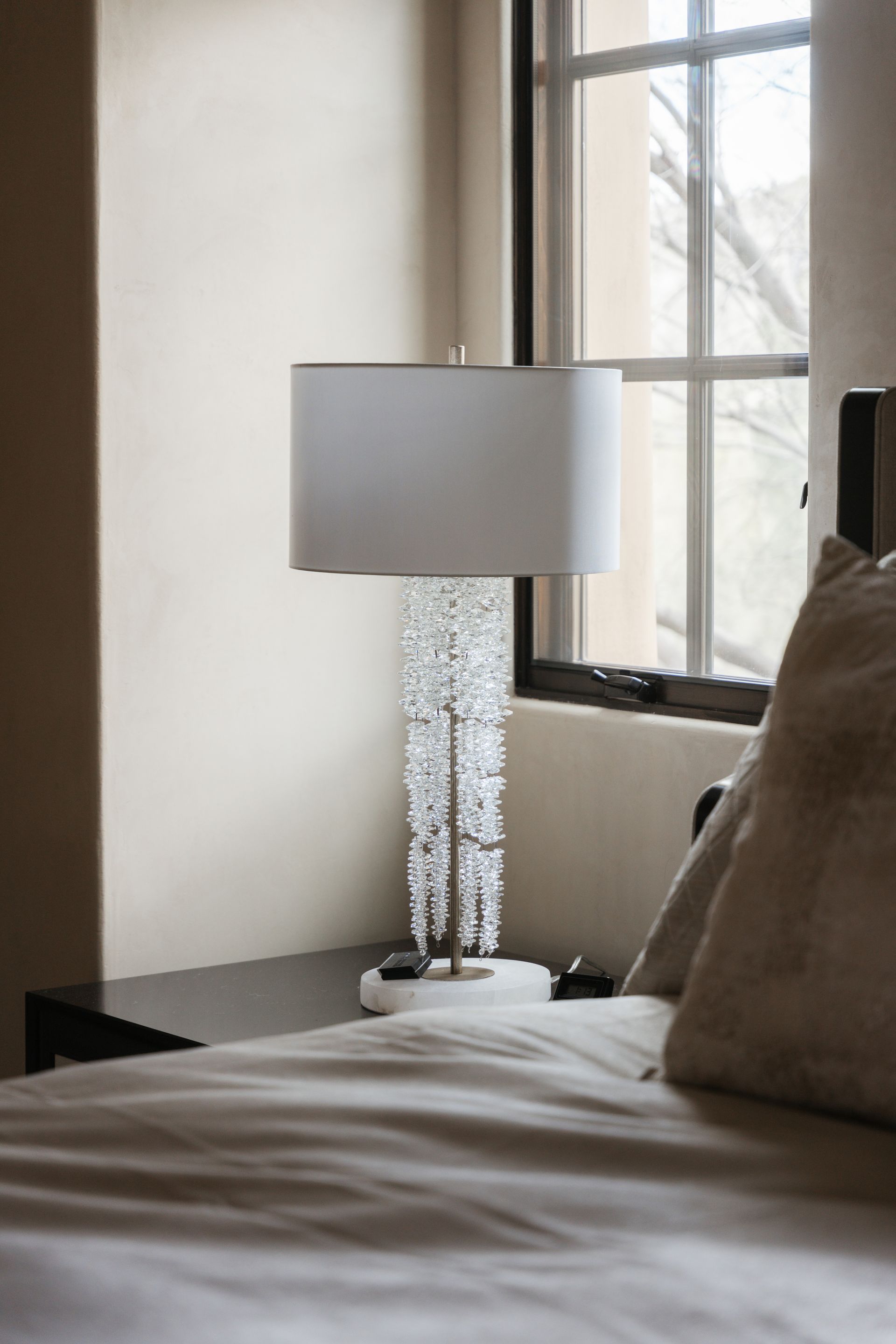 Bedside lamp with crystal embellishments on a marble base, near a window, bed in foreground.