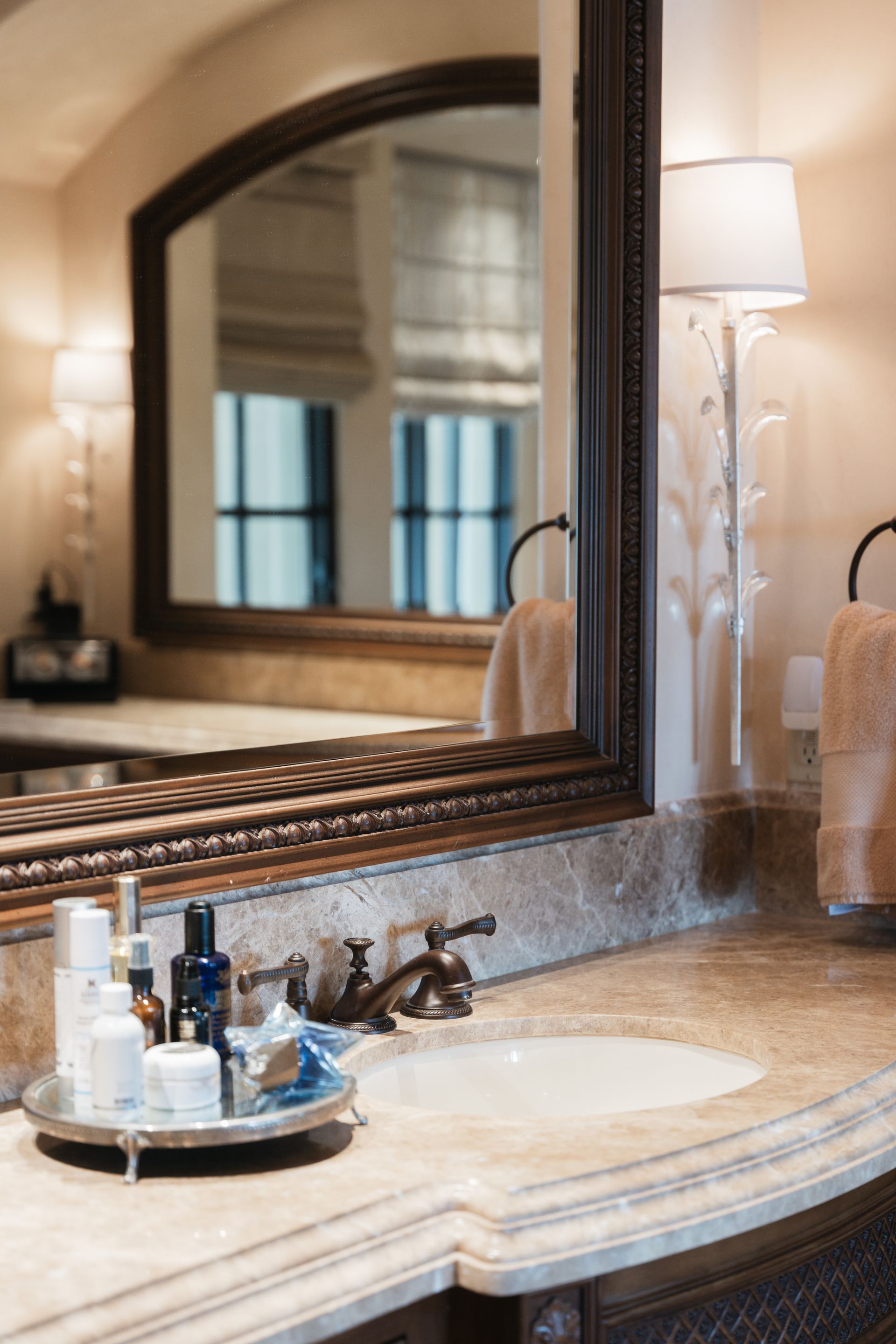 Bathroom vanity with ornate mirror, sink, skincare products, and wall sconces. Beige and brown color scheme.