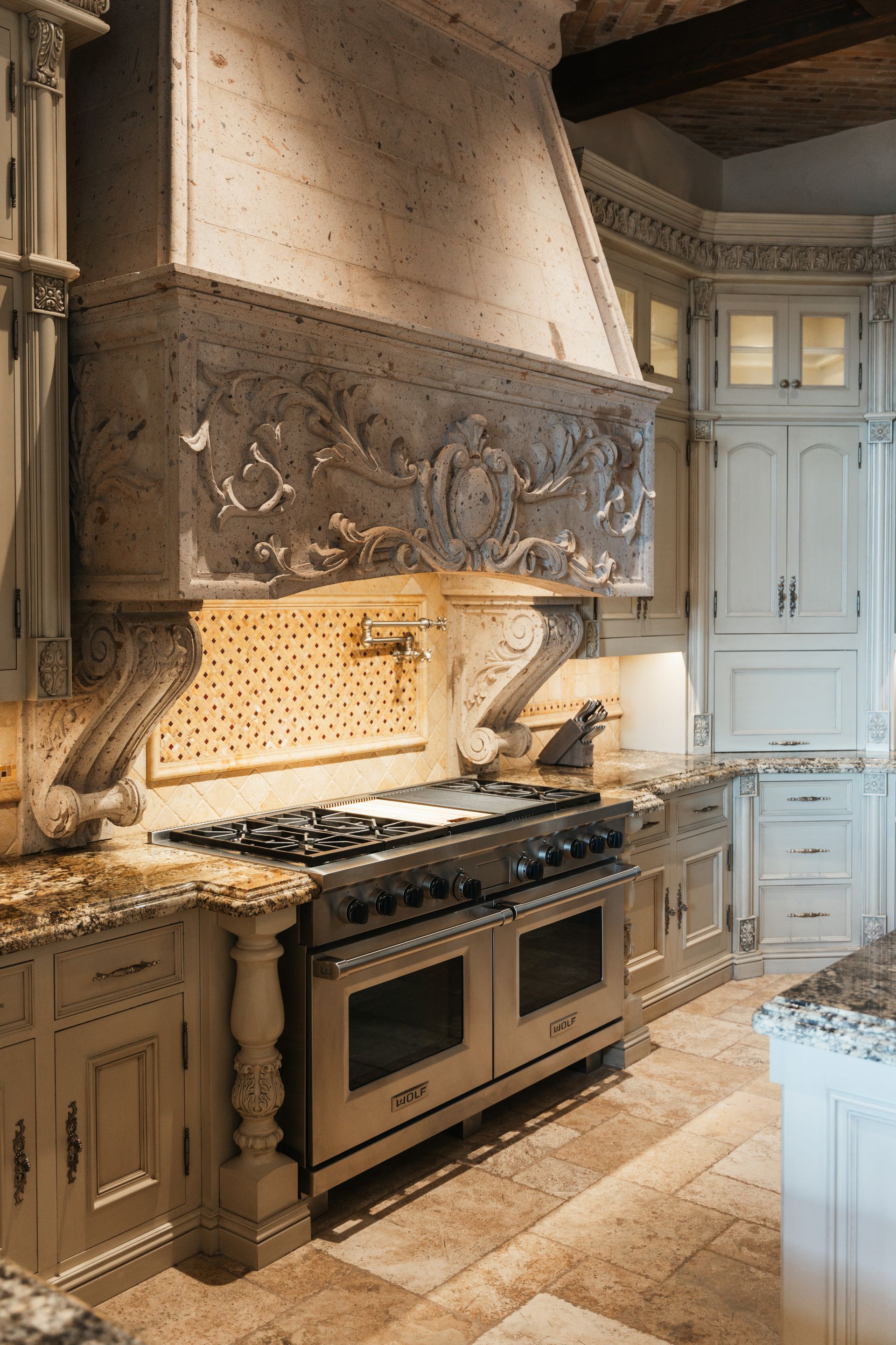 Kitchen with a large stainless steel range, ornate carved range hood, and light-colored cabinetry.
