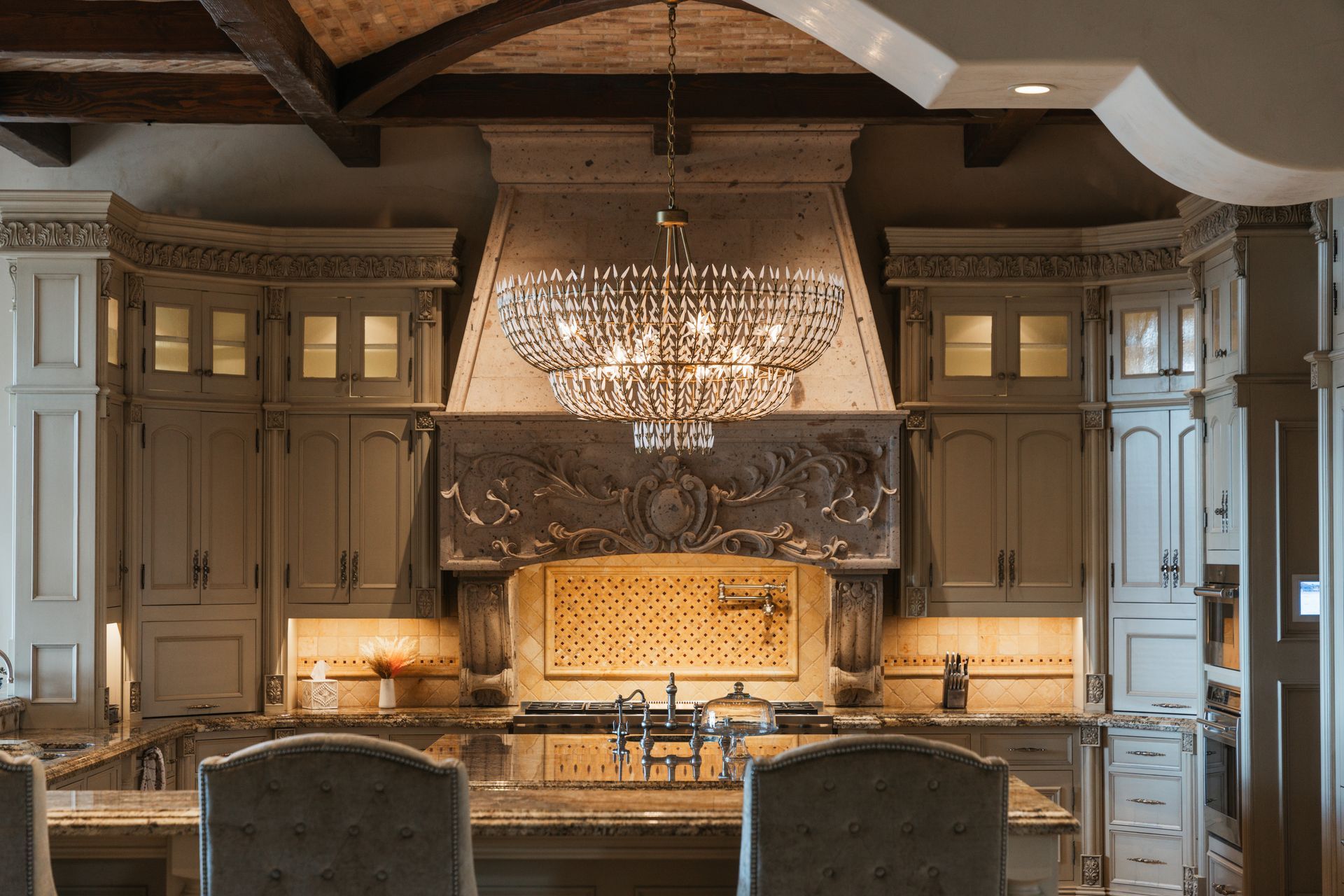 Elegant kitchen with ornate cabinetry, a large hood, and a sparkling chandelier.