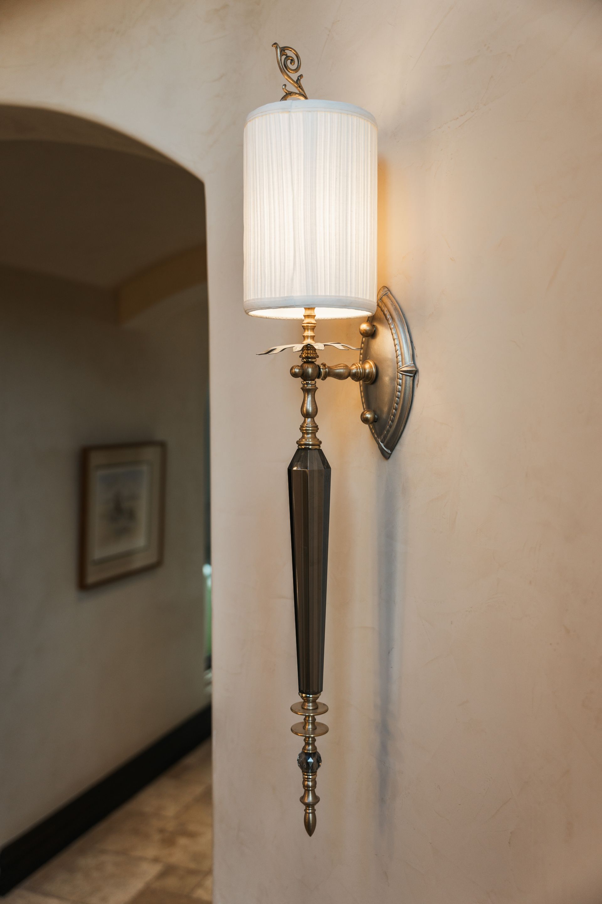 Wall-mounted light fixture with a lampshade. Beige wall, arched doorway, and a framed picture are in the background.