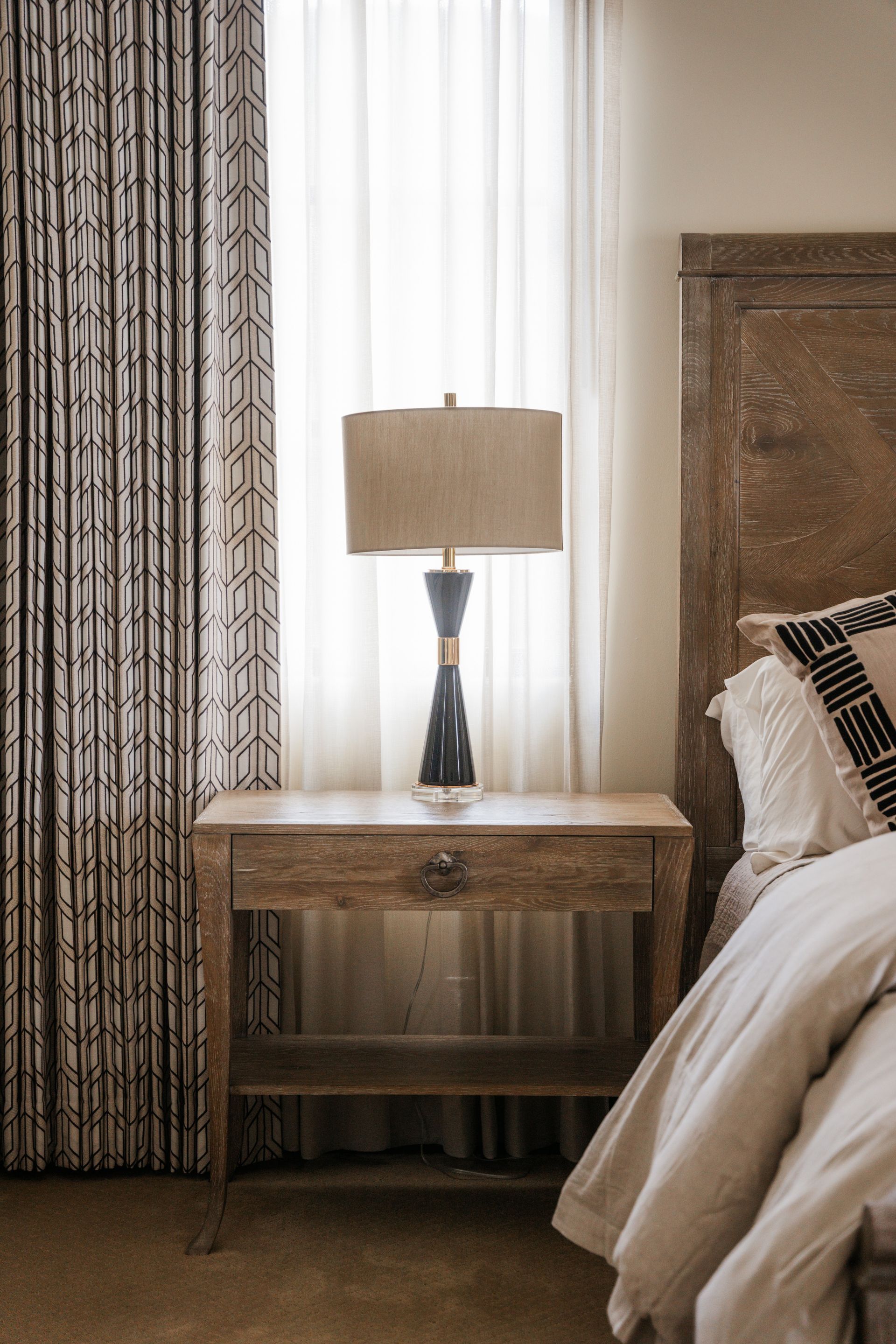 Bedroom nightstand with lamp, patterned curtain, sheer curtain, and headboard.