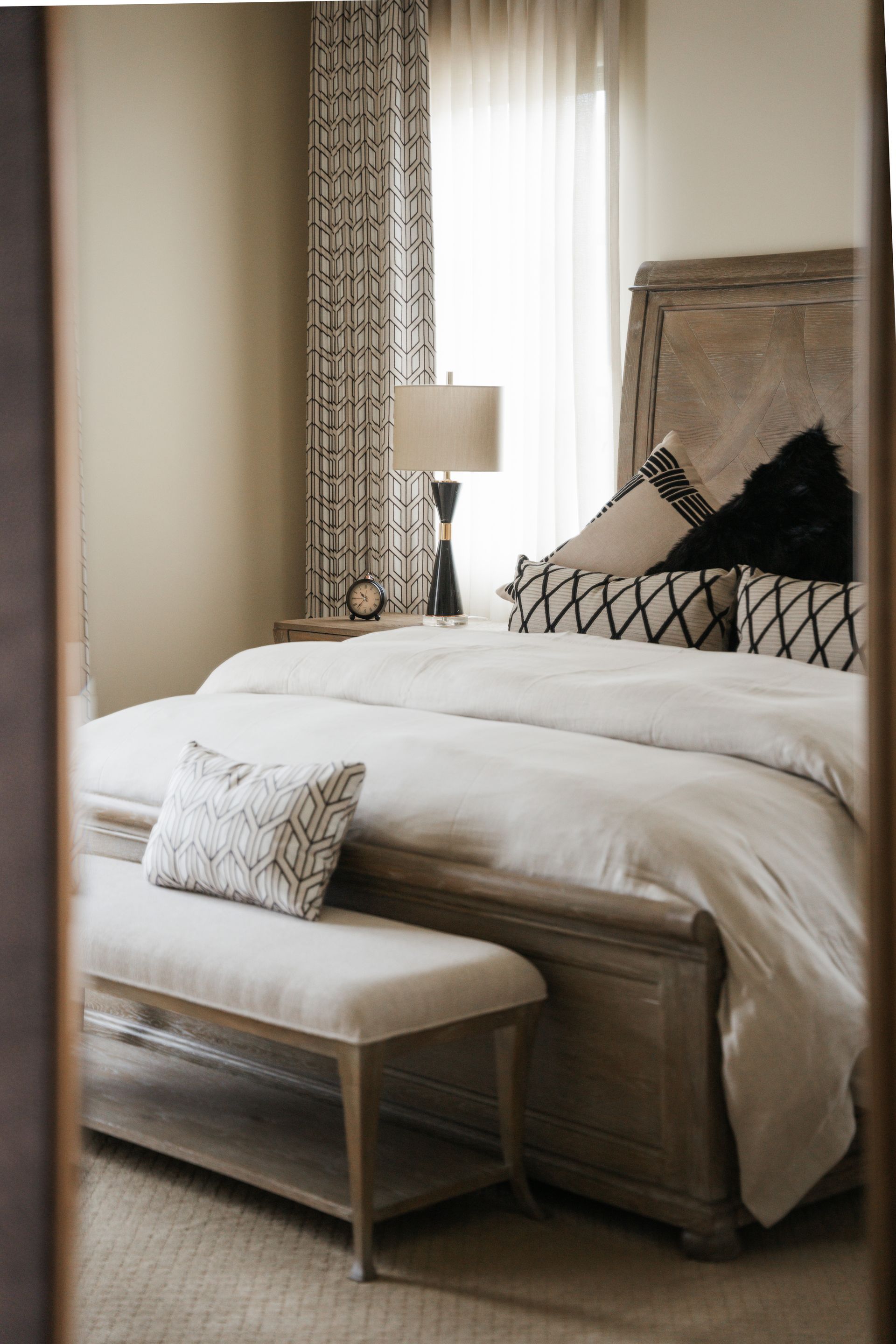 Bedroom with a wooden bed, patterned curtains, and neutral-toned bedding. A bench sits at the foot of the bed.