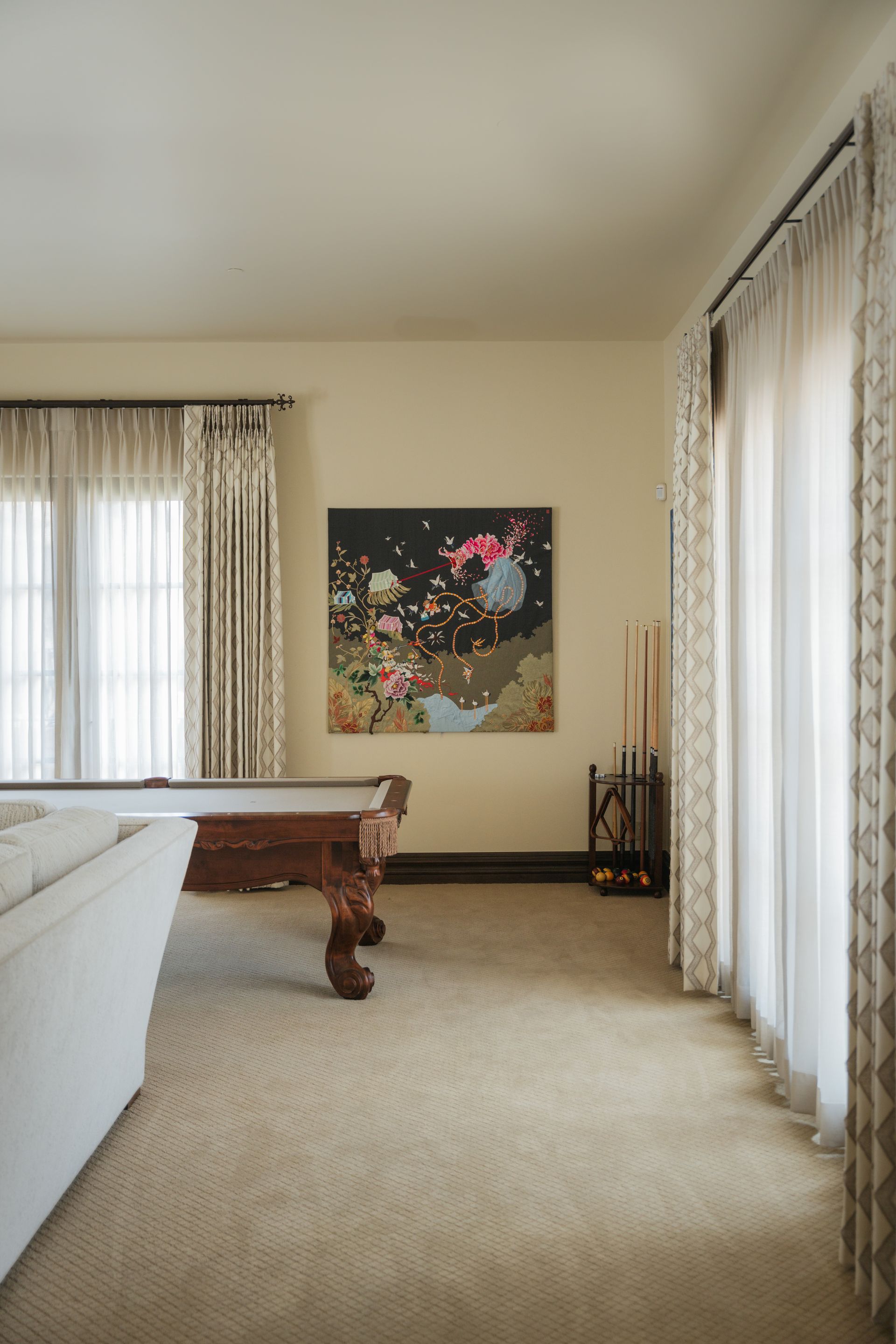 Pool room with pool table, artwork, and sheer curtains. Beige carpet and walls.