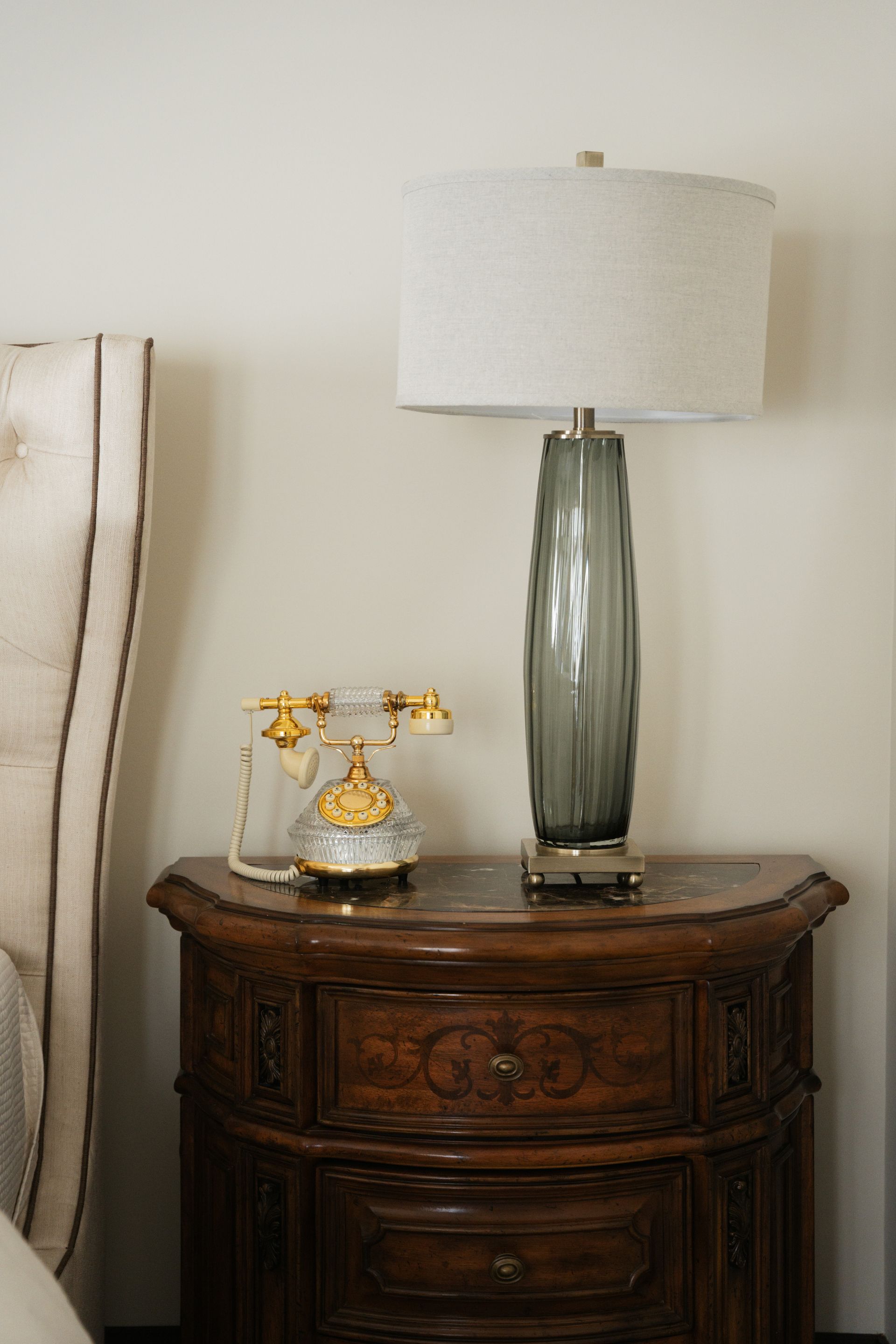 Bedroom nightstand with lamp, decorative phone, and headboard. Dark wood, gray lamp, neutral walls.