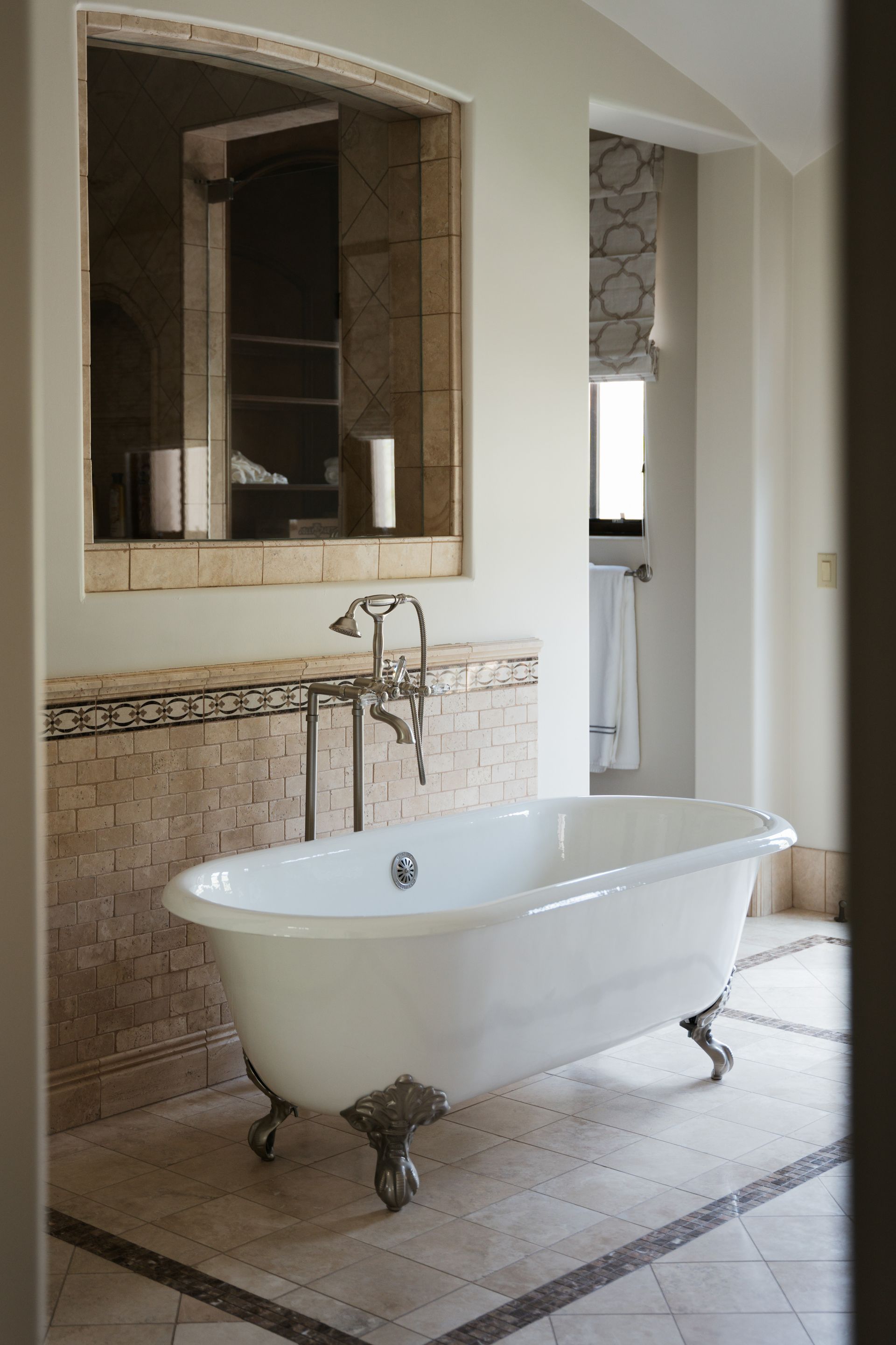 Claw-foot bathtub in a luxurious bathroom with a mosaic tile floor, mirror, and window with patterned blinds.