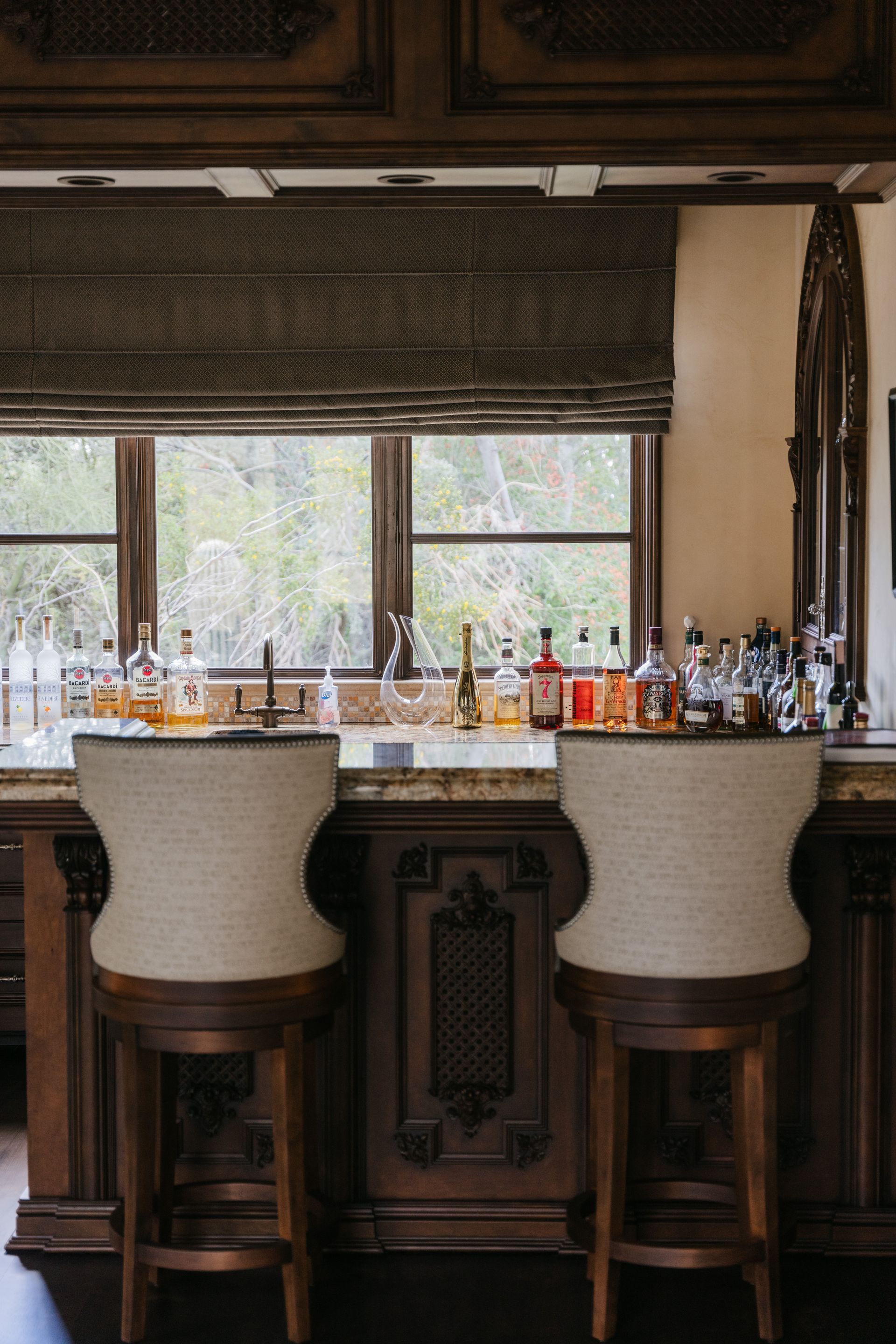 Two bar stools face a bar with bottles, window behind. Dark wood cabinets.