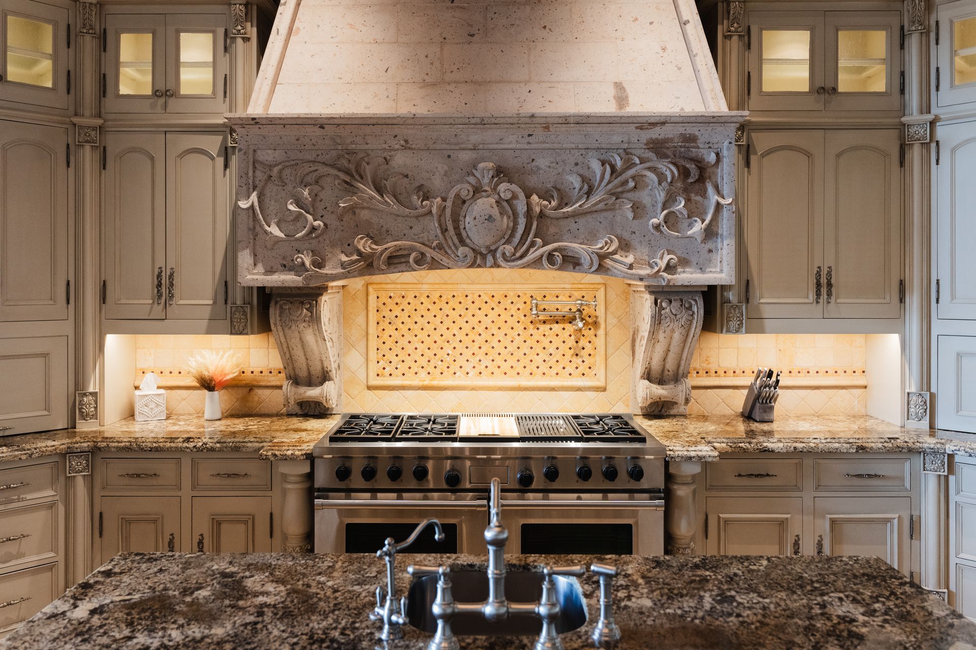 Kitchen with ornate, light-colored cabinets and range hood. Stainless steel stove and dark countertop.