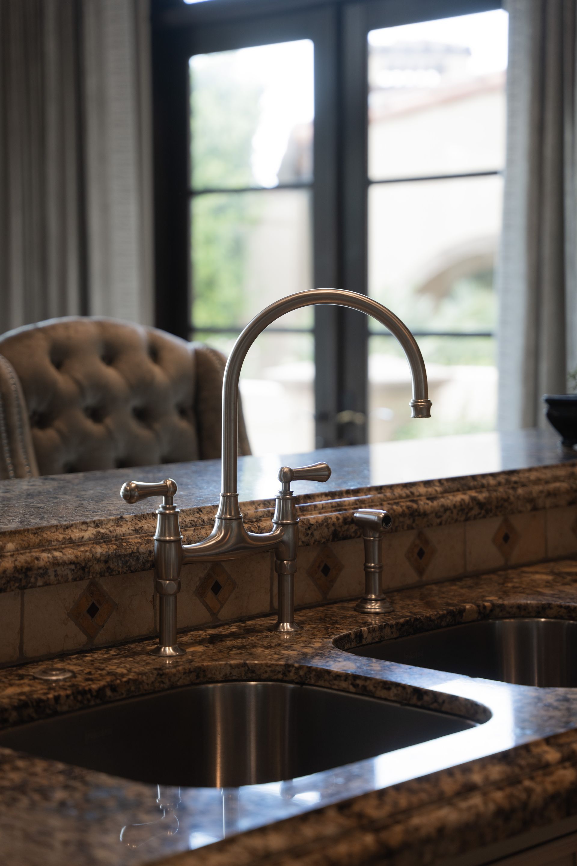 Kitchen sink with chrome faucet on a granite countertop, view of a window and upholstered chair in the background.