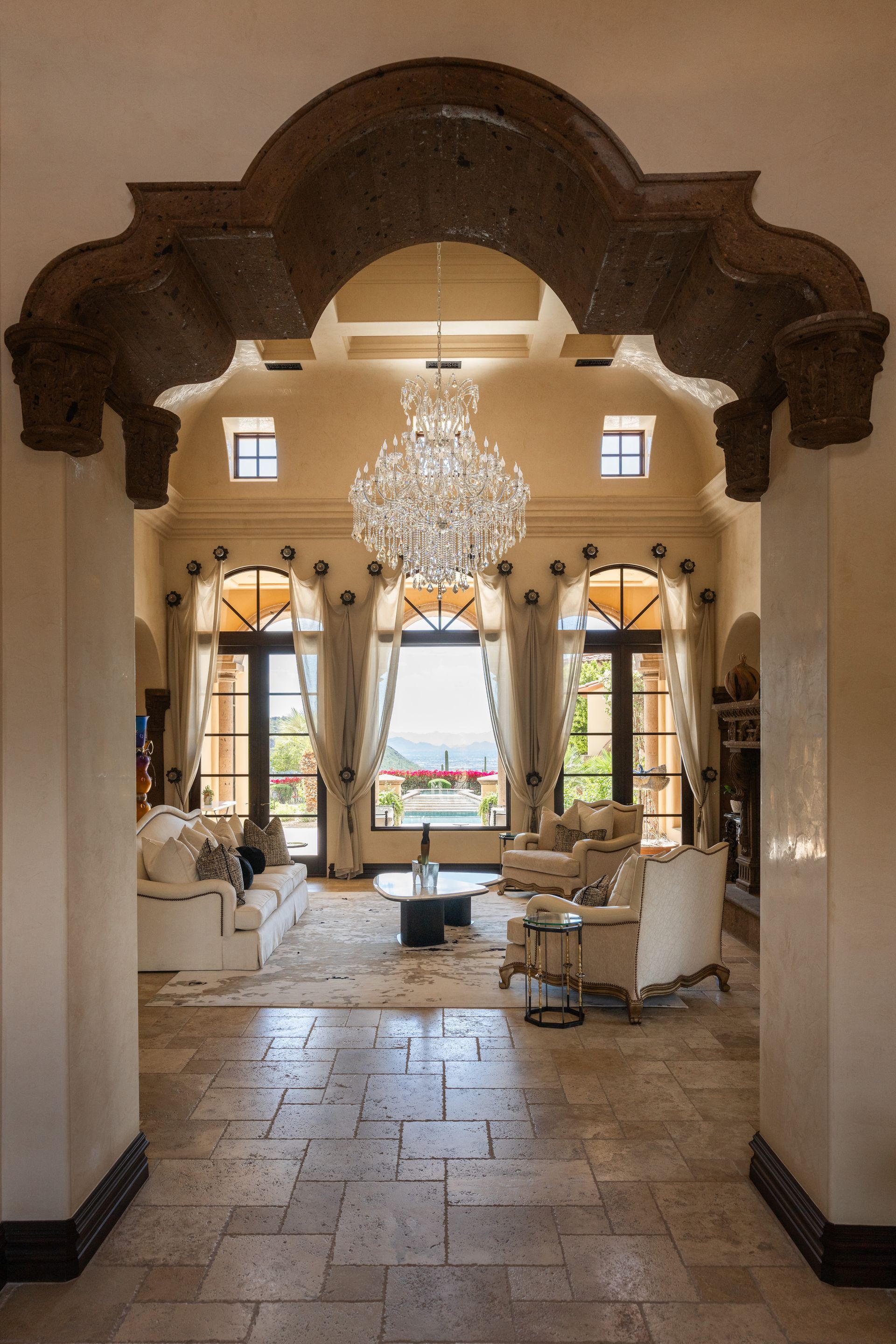Elegant living room, viewed through an arched doorway, with chandelier, beige walls, and views of a patio and mountain.