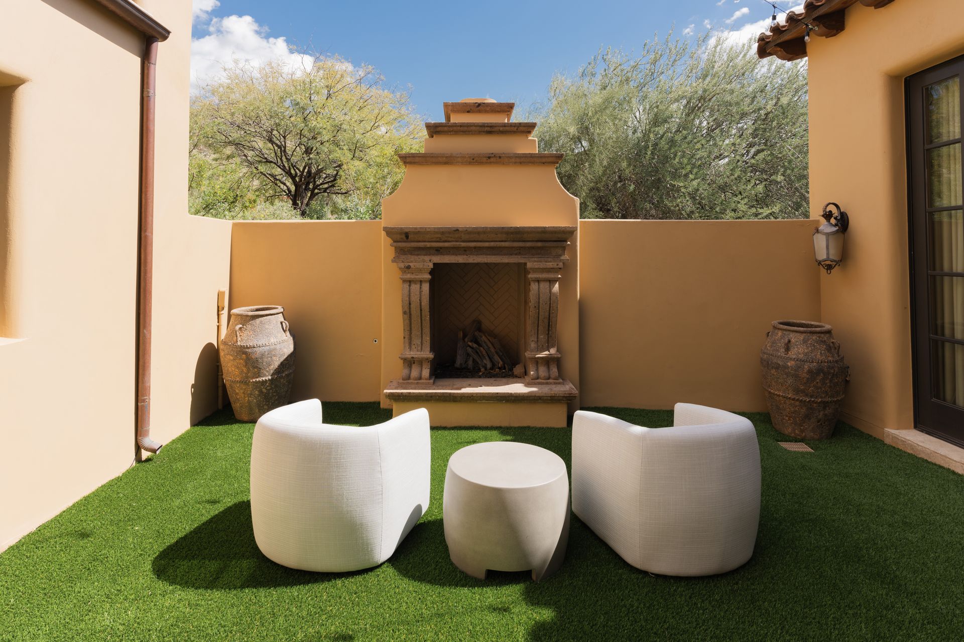 Patio with fireplace, two white chairs, and a small table on green turf, set against tan walls.