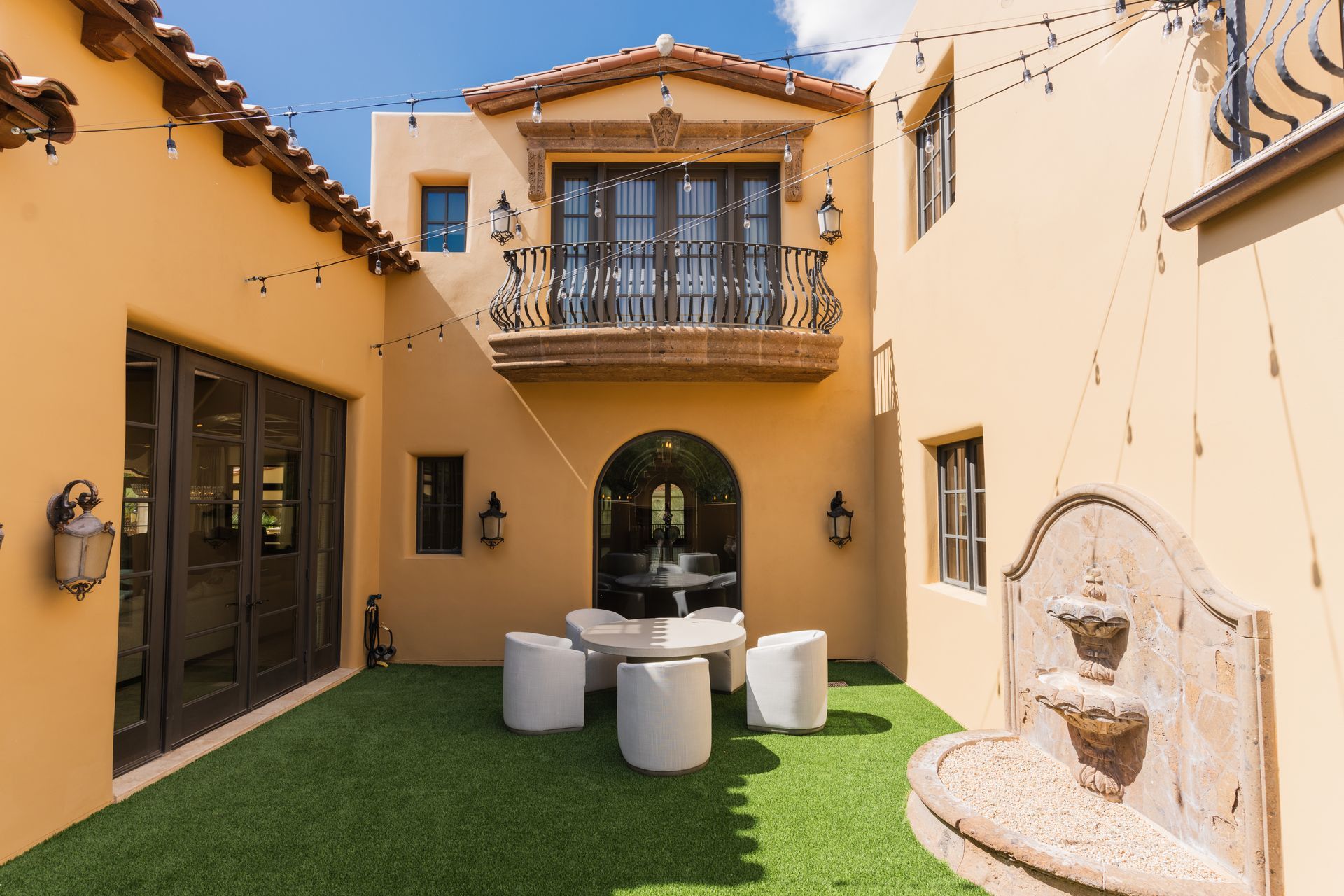 Courtyard with faux grass, fountain, patio set, and balcony. Tan stucco walls and string lights.