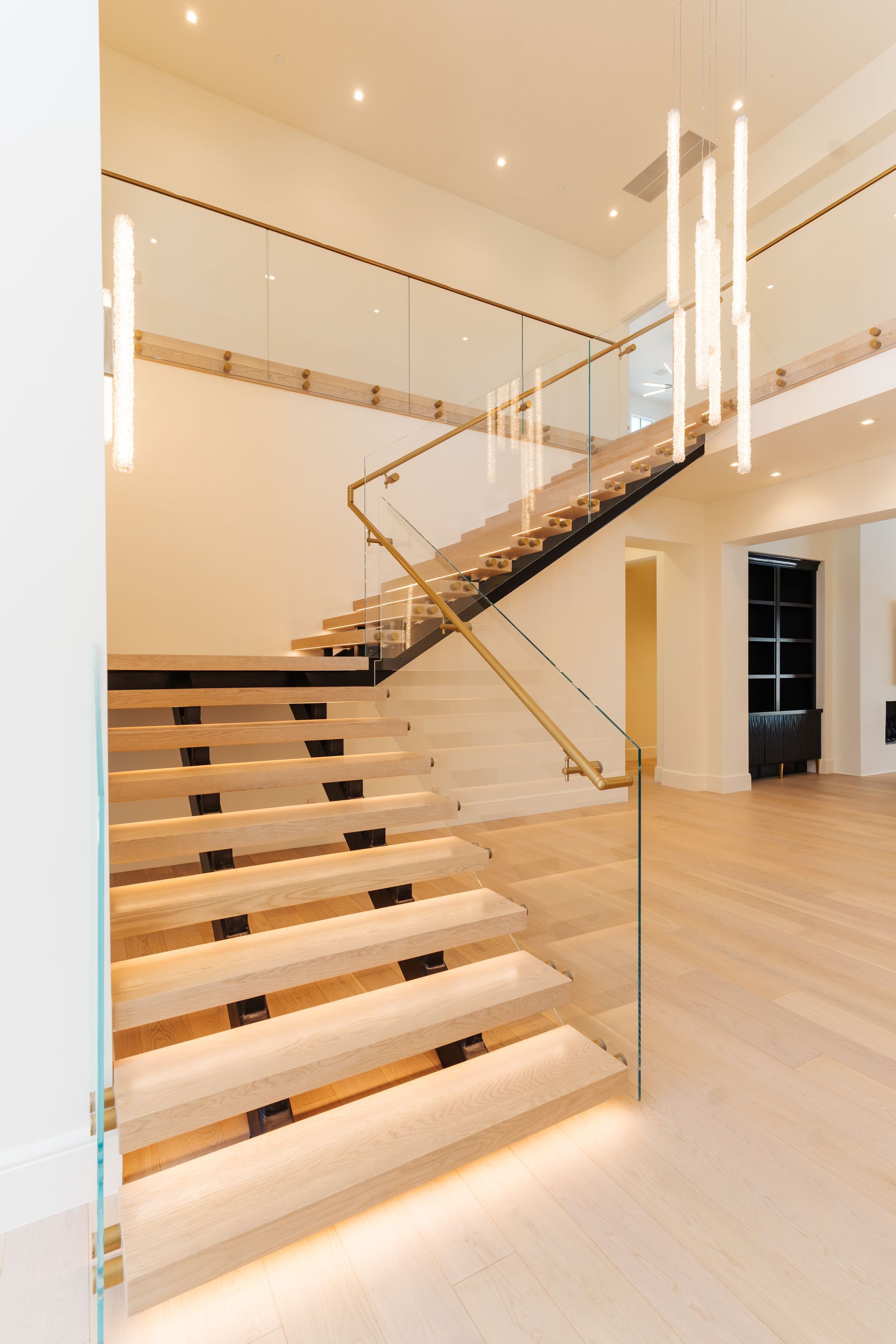 Modern staircase with wood steps, glass railing, and gold accents. Bright, open entryway with hanging lights.