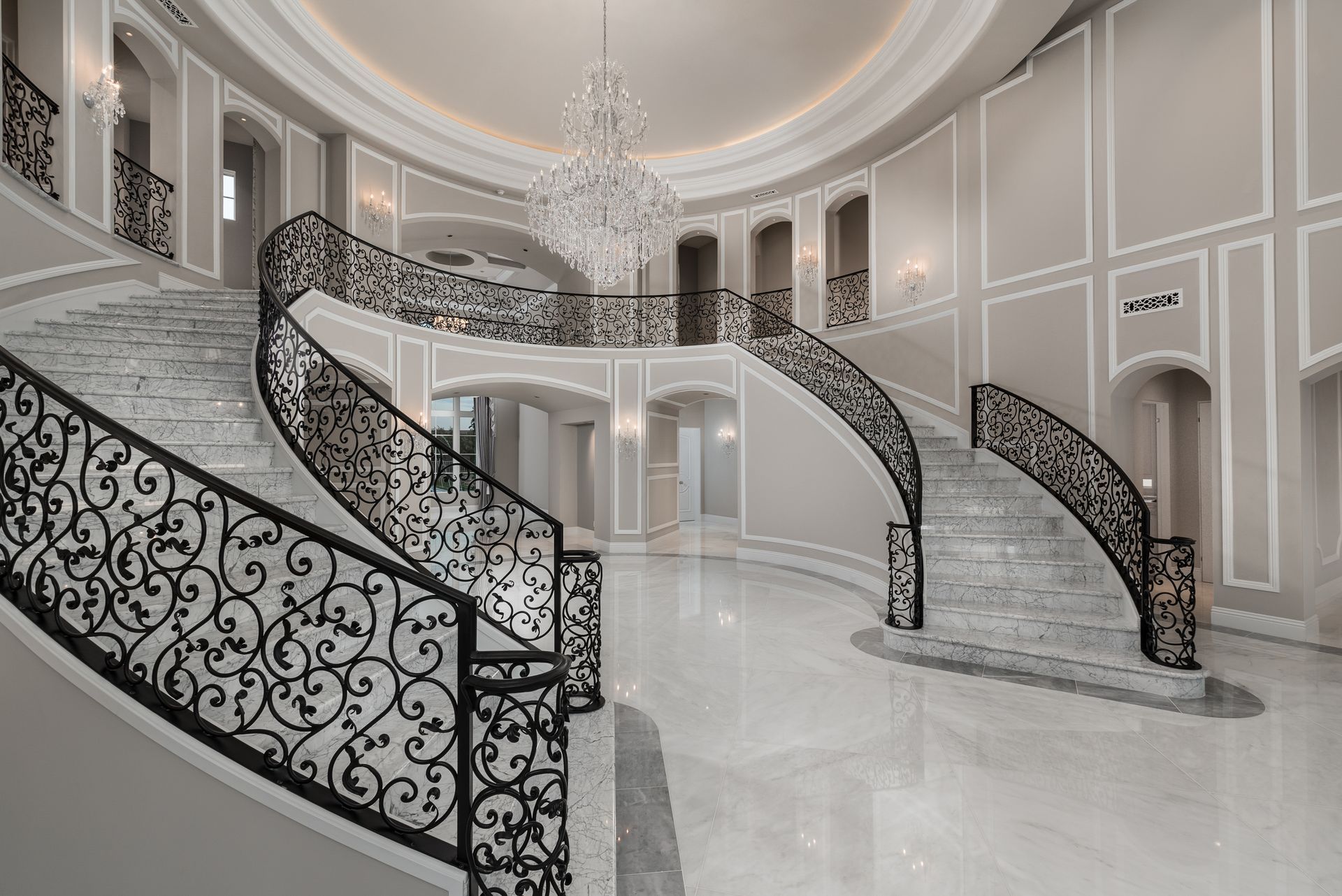 Grand foyer with curved staircases, marble floors, ornate black railings, and a chandelier.