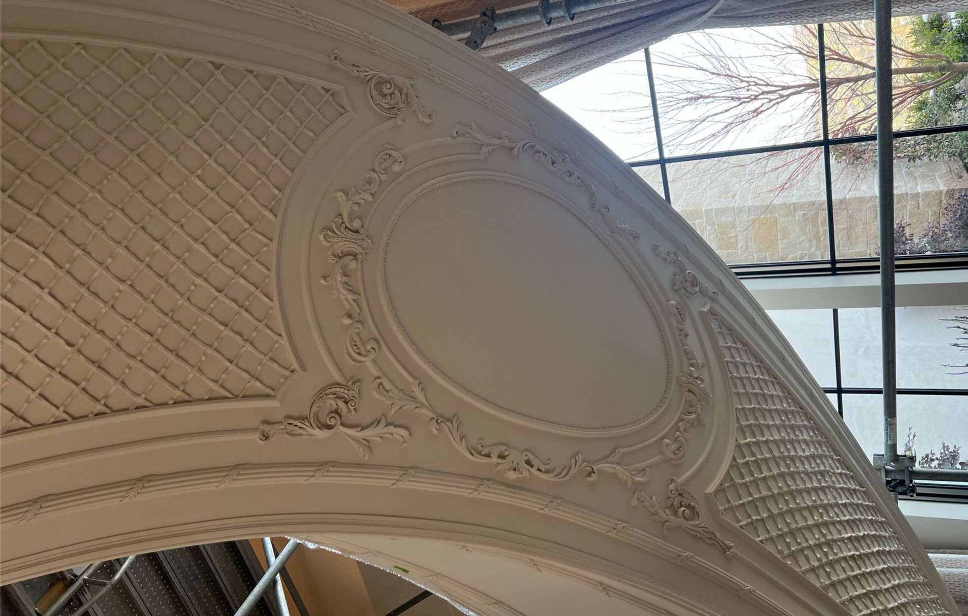 Ornate, white ceiling with oval molding and lattice pattern. Sunlight streams through a nearby window.