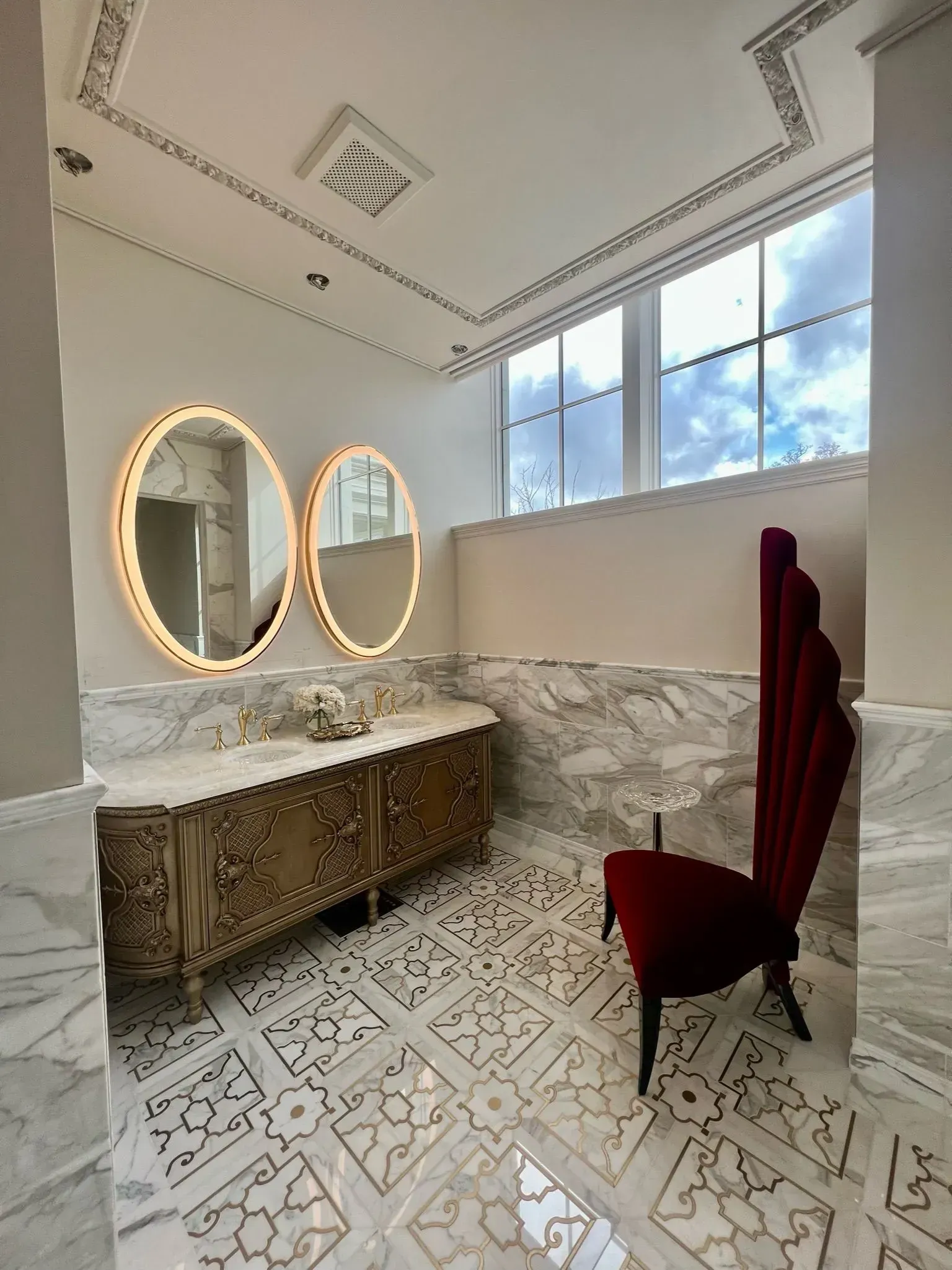 Ornate bathroom with gold cabinet, oval mirrors, and high-backed red chair. Marble and patterned tile floors.