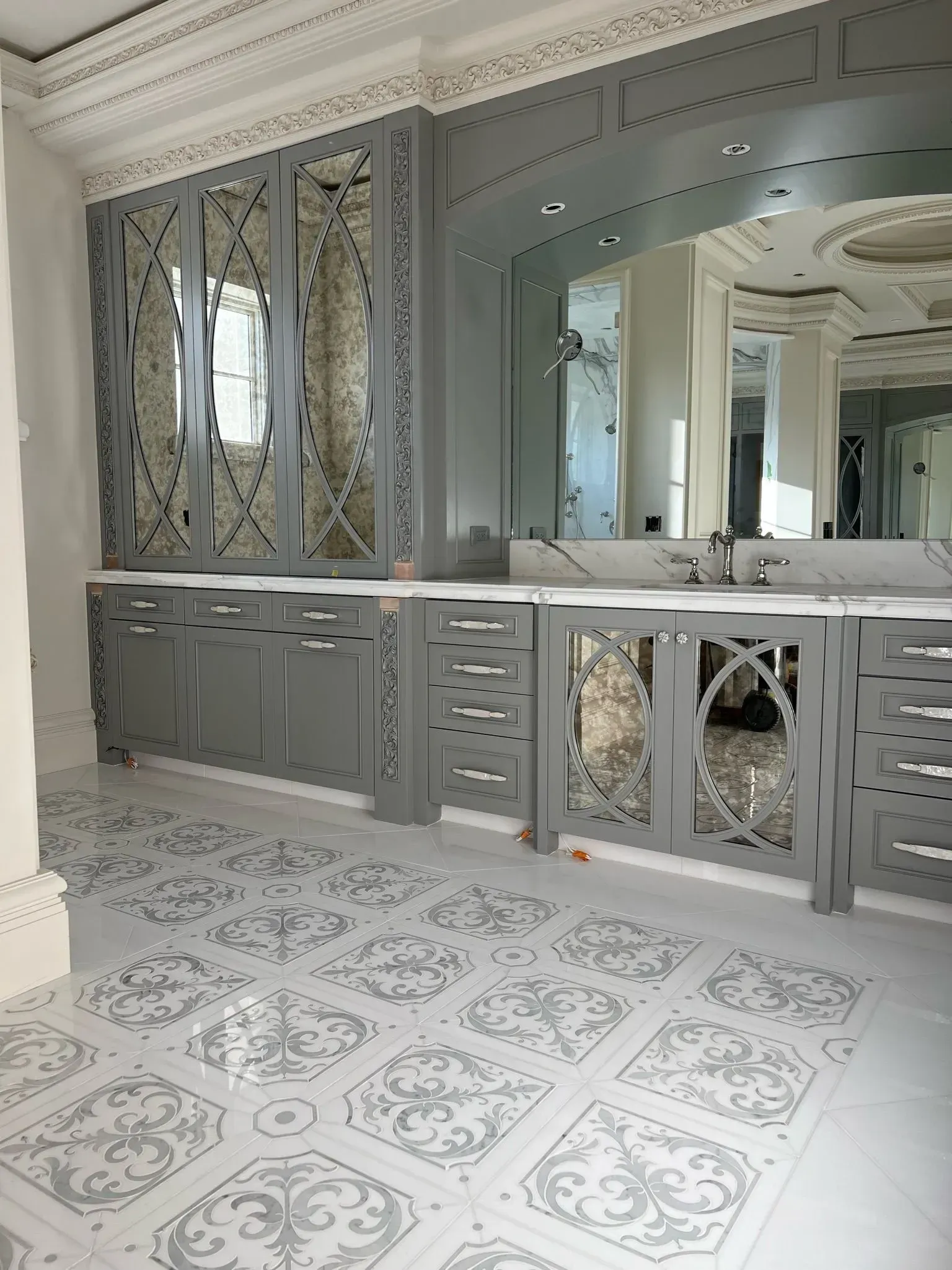 Gray and white bathroom with intricate mirror-backed cabinets, ornate patterned floor, and large mirror.