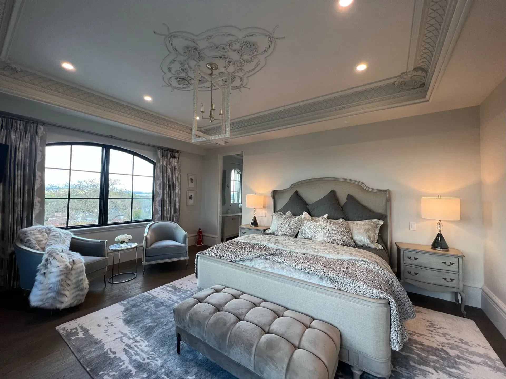 Luxurious bedroom with gray and white decor, large bed, seating area by window, and ornate ceiling.