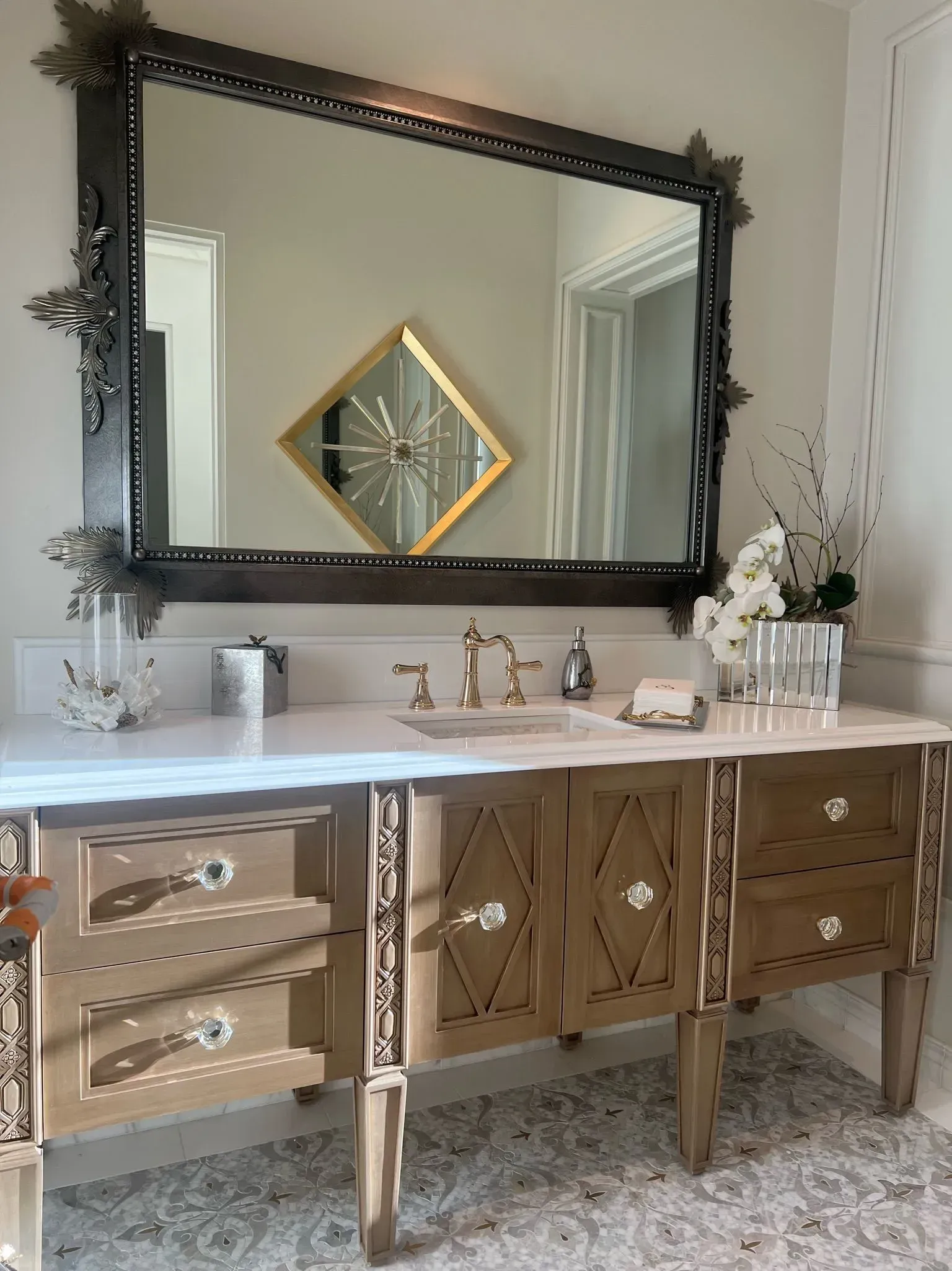Bathroom vanity with large decorative mirror, light wood cabinetry, white countertop, and gold fixtures.