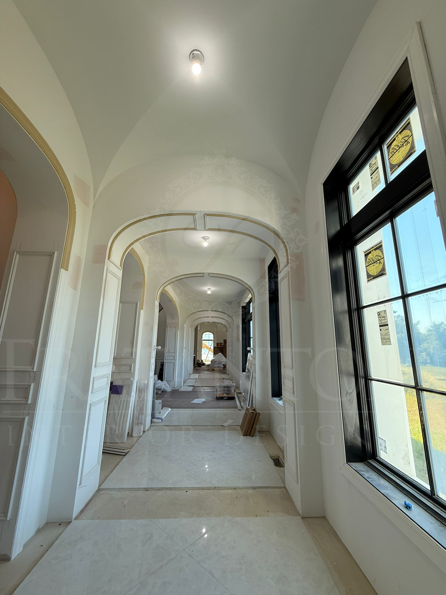 A sunlit hallway under construction, featuring multiple white arched doorways and large black-framed windows.