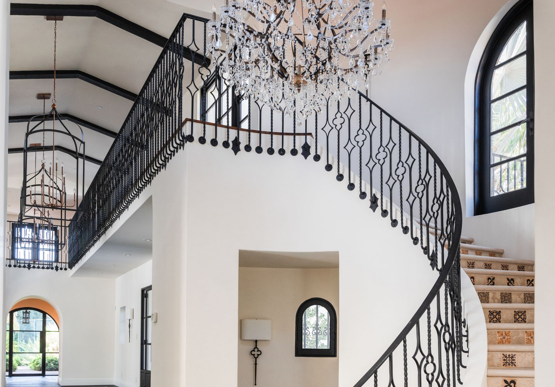 A grand foyer with a curving black wrought-iron staircase, white walls, and a large chandelier.