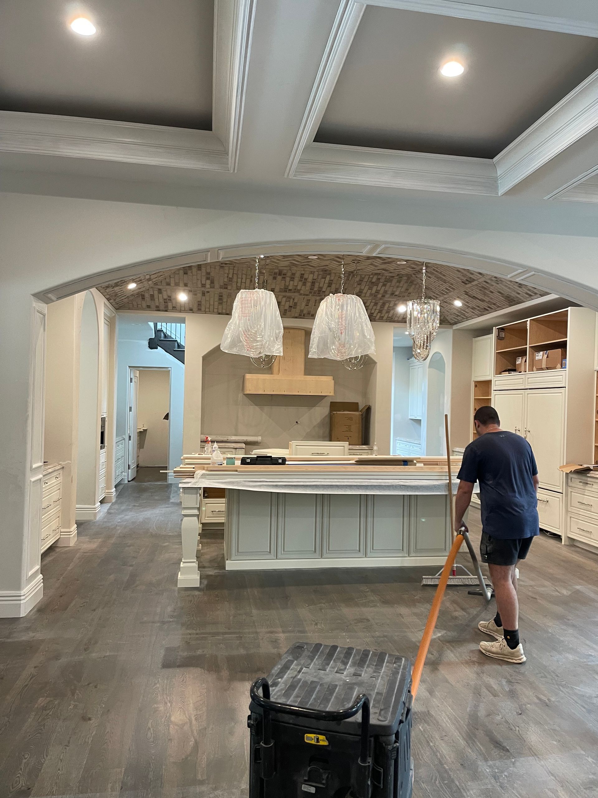 Man sweeping a large kitchen under construction. Gray floor, light cabinets, island.