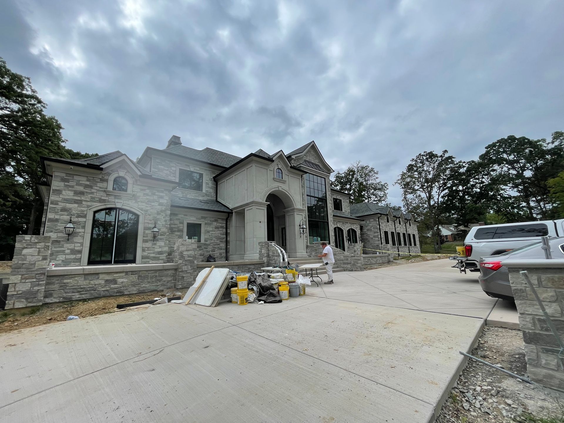 Large stone house under construction, concrete driveway, overcast sky.