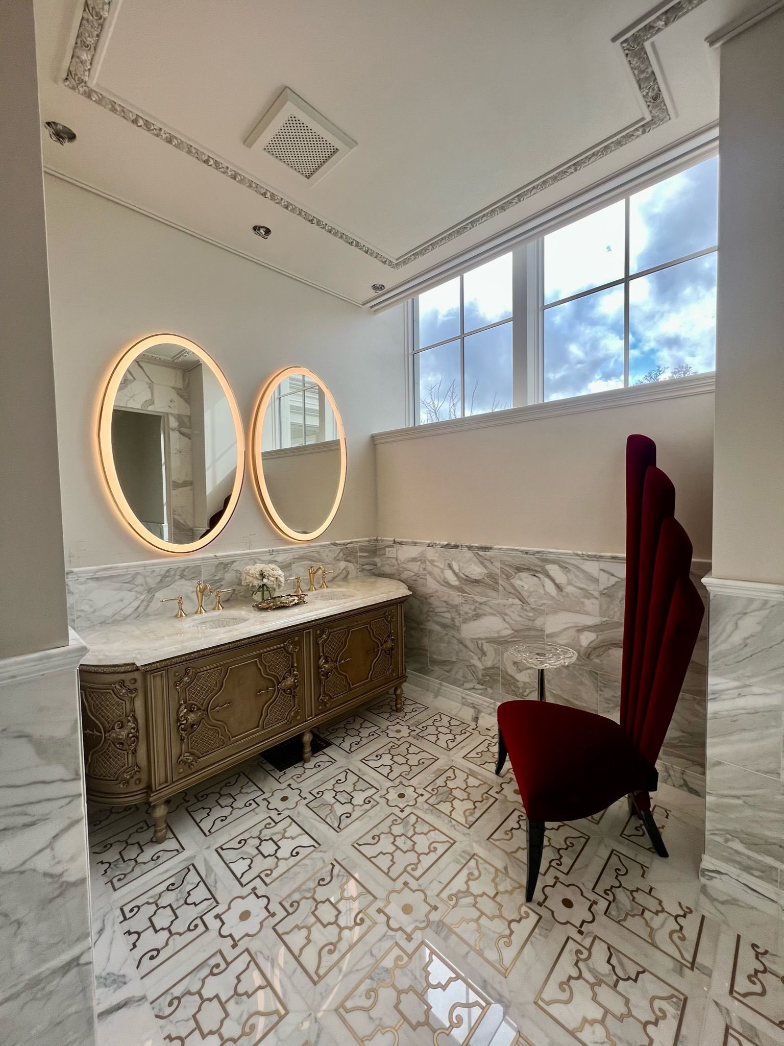 Elegant bathroom with ornate gold cabinet, oval mirrors, and burgundy velvet chair.