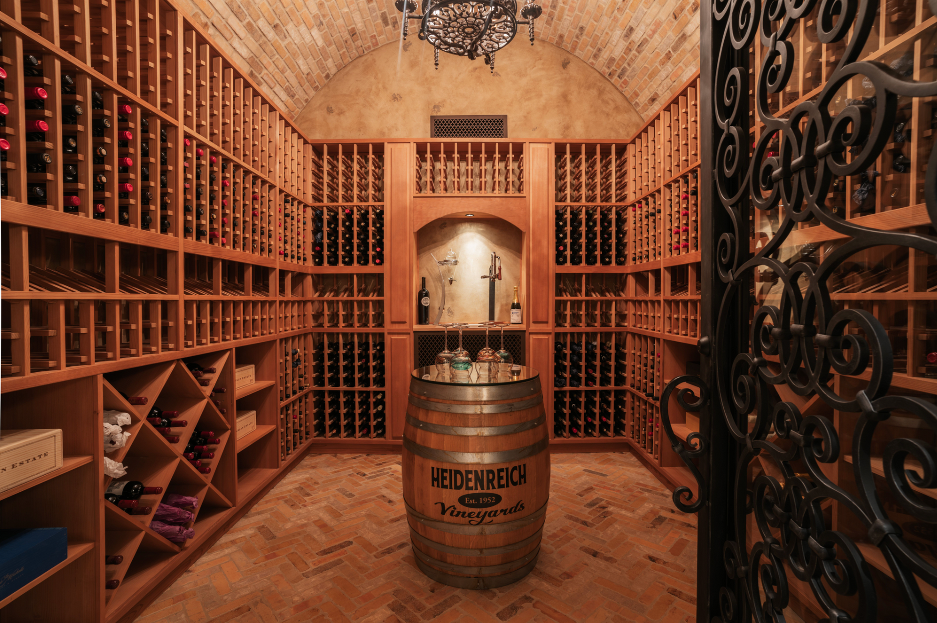 Custom luxury wine cellar with wooden racks filled with bottles, arched ceiling, barrel, and ornate iron gate.
