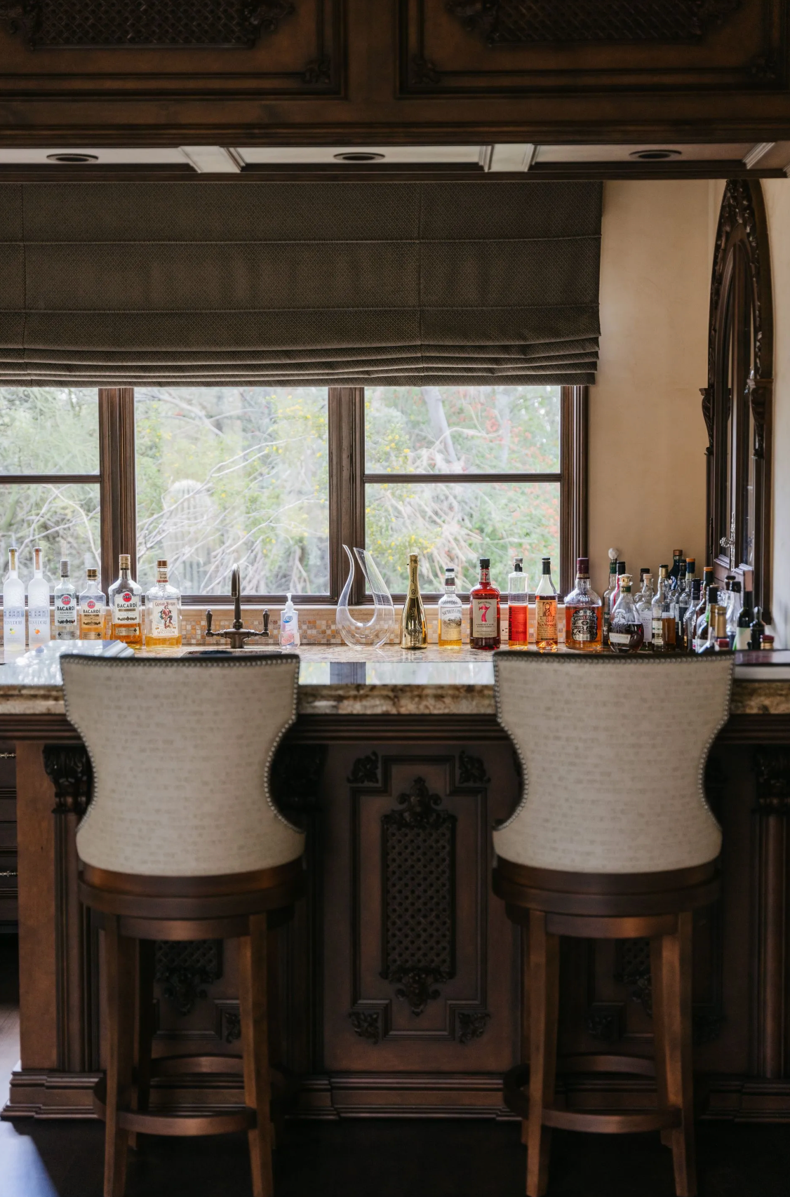 Two upholstered bar stools face a bar with bottles, under a window with a shade.