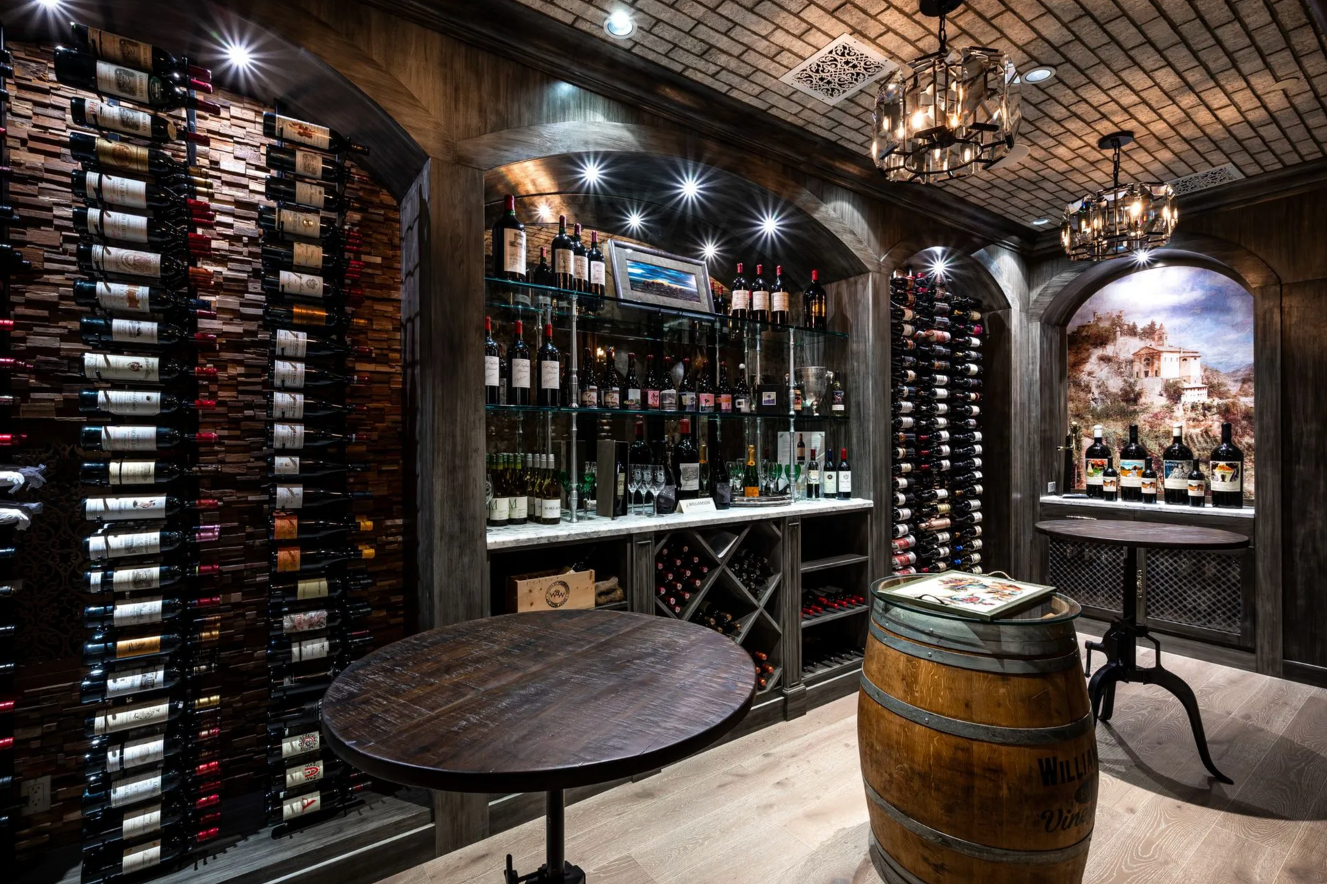 Wine cellar with arched walls, filled with bottles; a table and barrel sit in the foreground, lit by chandeliers.