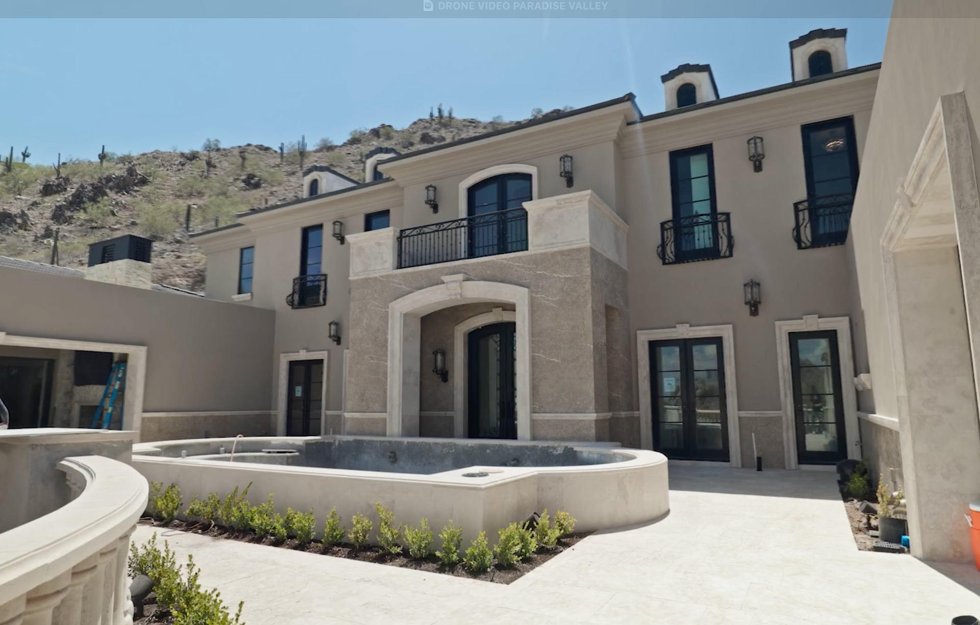 Luxury home exterior, light tan stucco, arched entrance, multiple balconies, mountain backdrop.