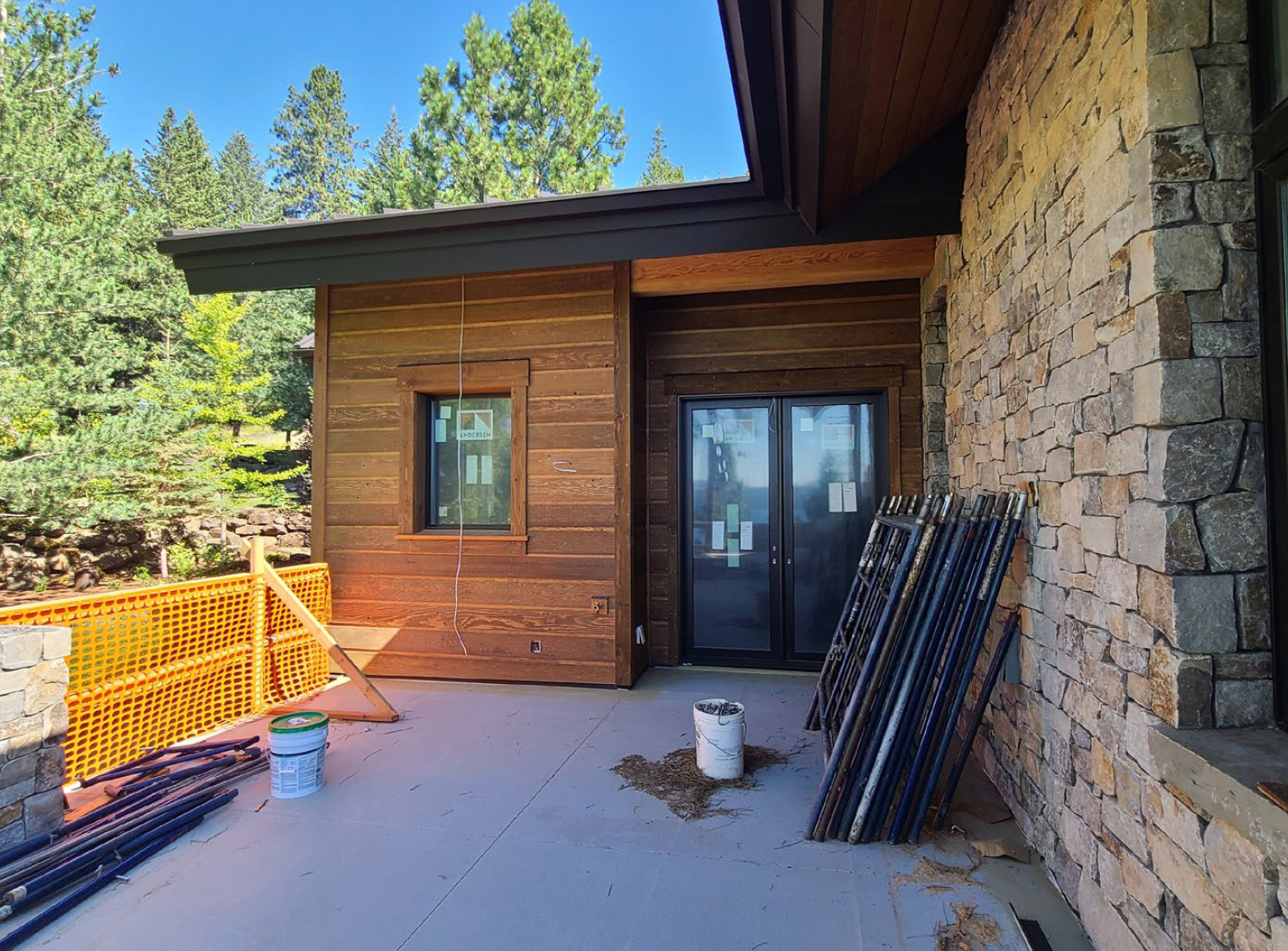 Exterior of a modern home under construction. Stone wall, wood siding, sliding glass door, and stacked materials are visible.