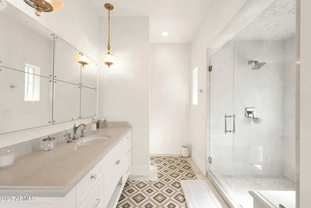 Bright white bathroom with marble countertop, glass shower, and patterned floor tiles.