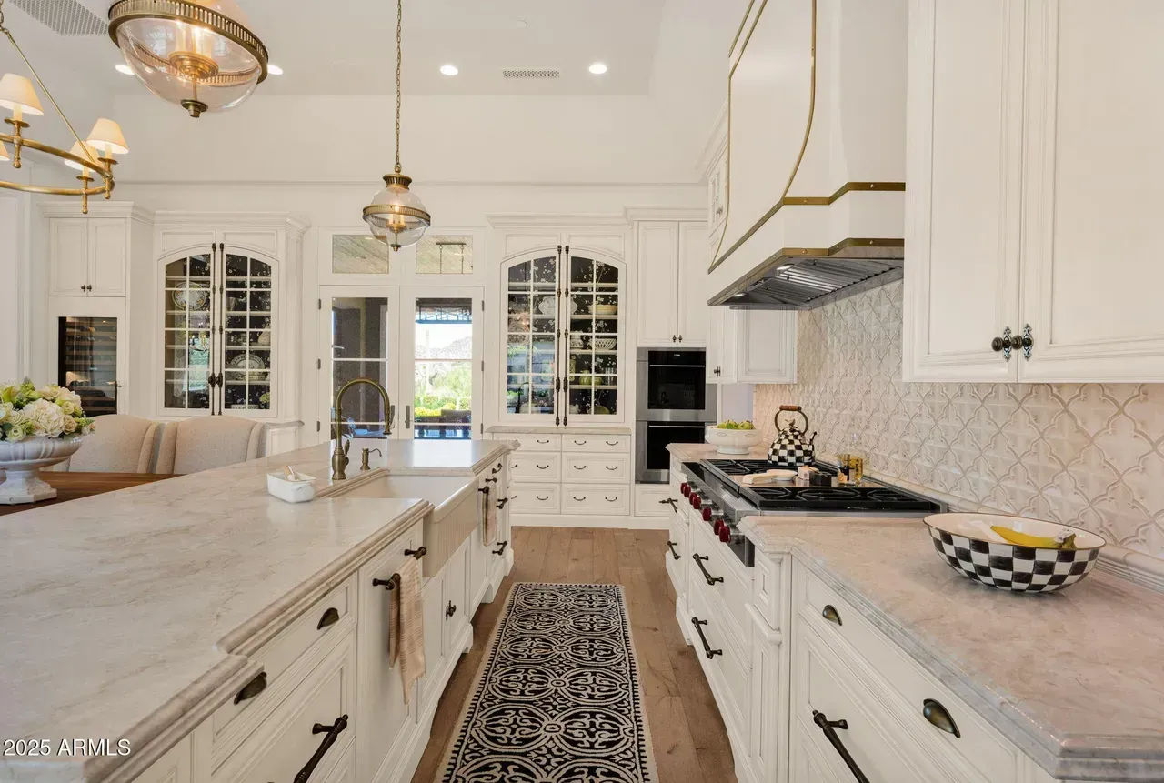 White kitchen with marble countertops, black and white rug, glass cabinets, and gold light fixtures.