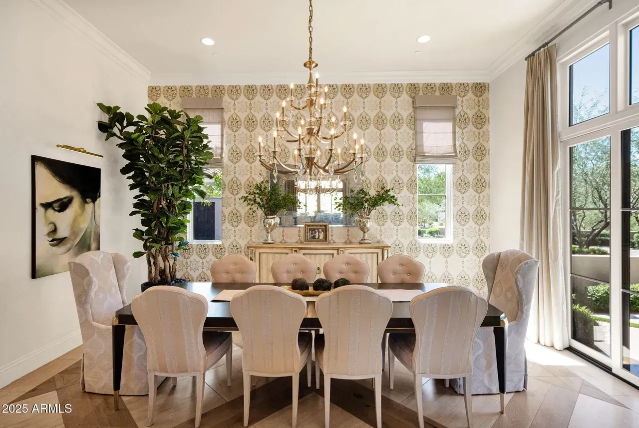 Elegant dining room with chandelier, long table, upholstered chairs, patterned wallpaper, and large windows.