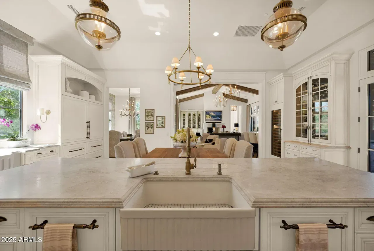 Elegant white kitchen with central island, sink, and view into dining area. Brass fixtures.