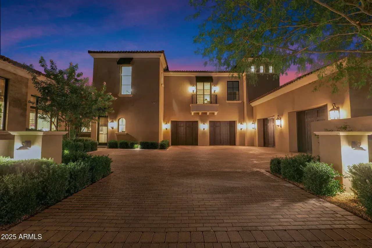 Elegant beige house with a brick driveway at dusk, with ambient lighting and bushes.