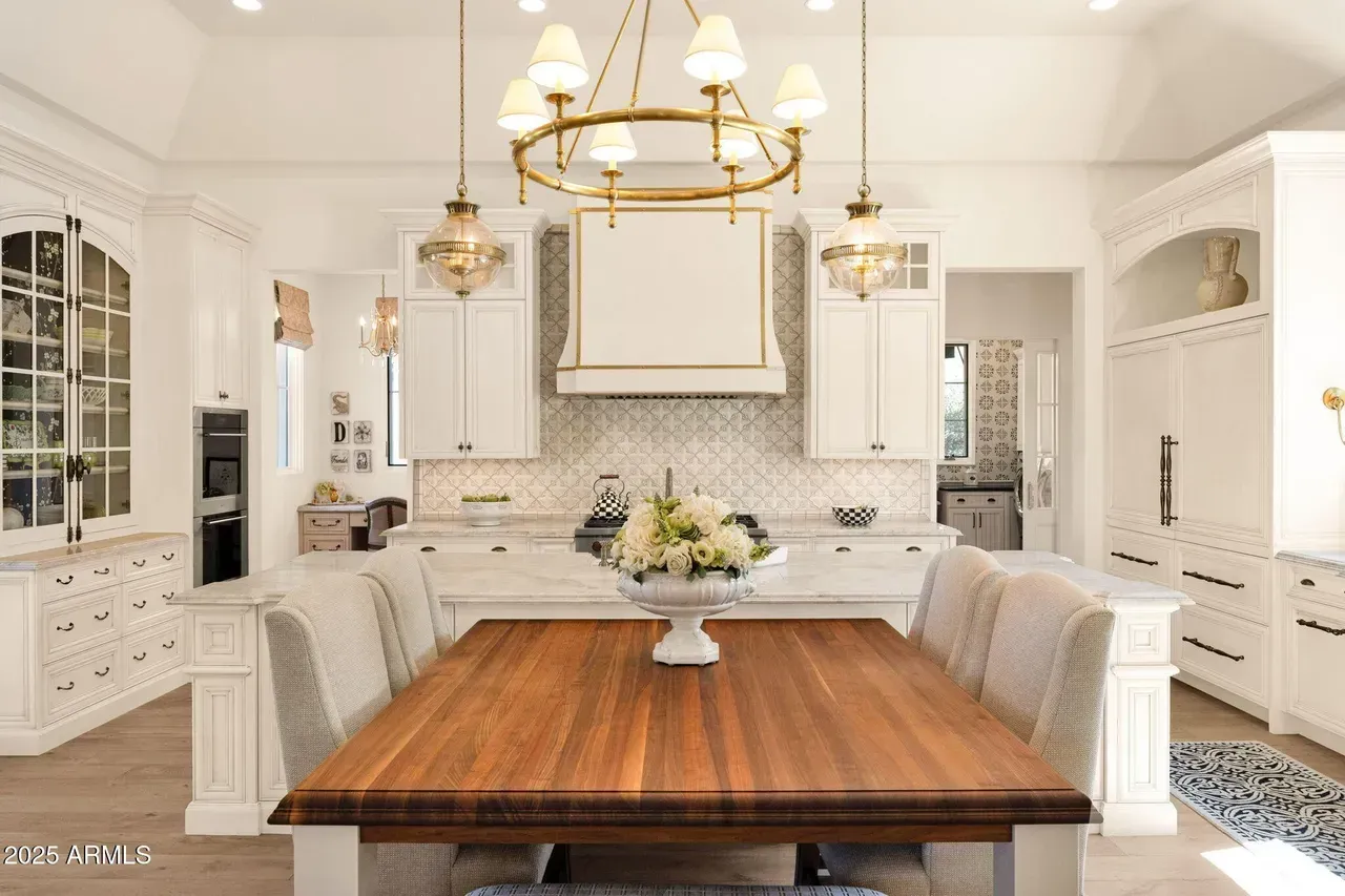 Bright white kitchen with wooden dining table, gold chandelier, white cabinets, and island.