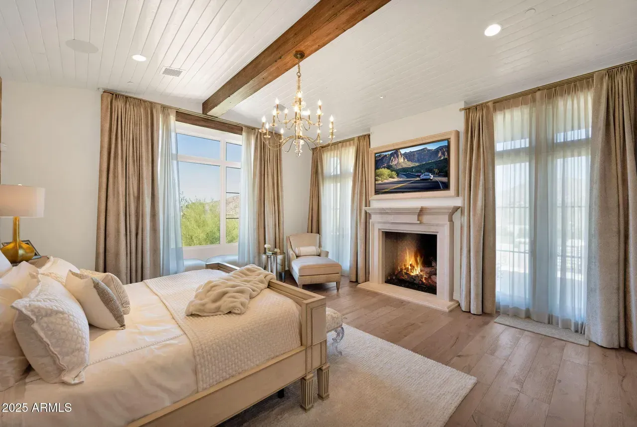 Bedroom with bed, fireplace, large windows with sheer curtains, chandelier, and wood beams.