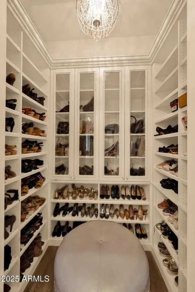 Walk-in closet with shelves of shoes and handbags; a central ottoman; and a crystal chandelier.