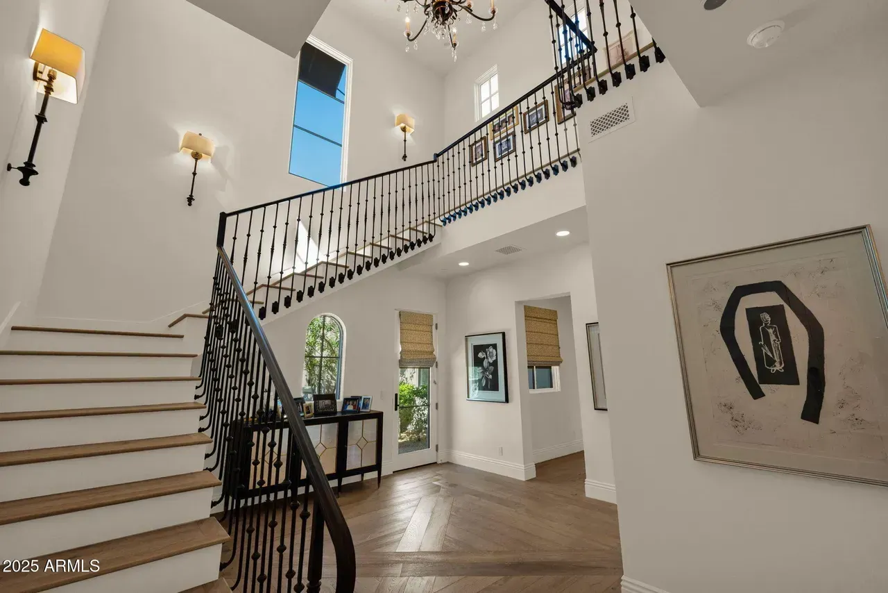Spacious white foyer with staircase, artwork, and chandelier. Natural light streams through windows.