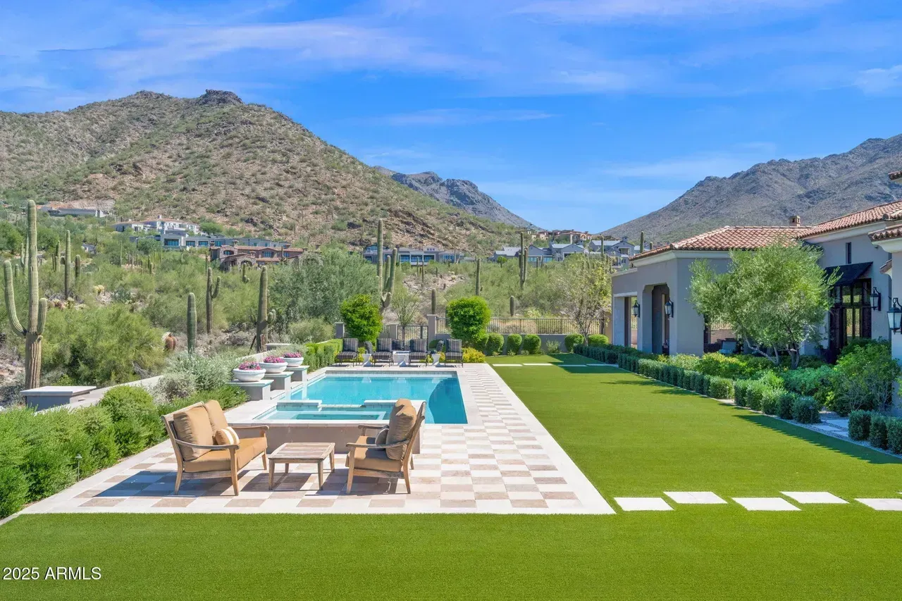 A pool and lawn with mountain views. Two chairs sit poolside. Bright blue sky overhead.