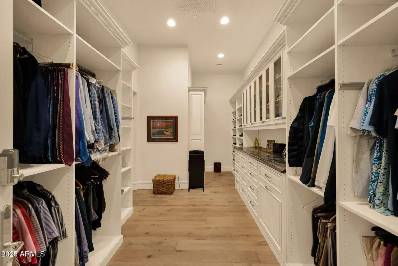 Luxury Walk-in closet with white shelves and clothing racks; light wood floor; small decor and cabinet space.