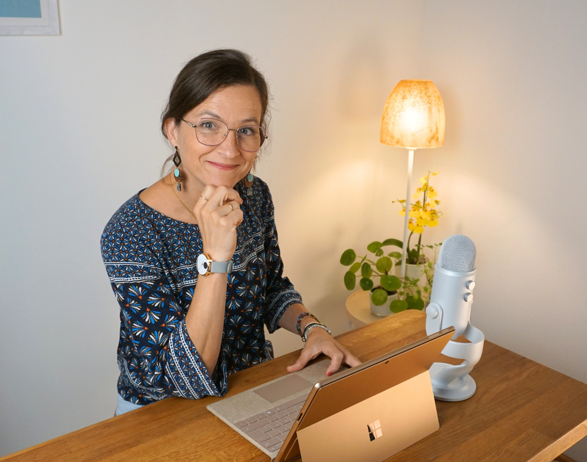Frau am Schreibtisch mit Laptop, Mikrofon und Lampe, lächelnd, die Hand am Kinn.