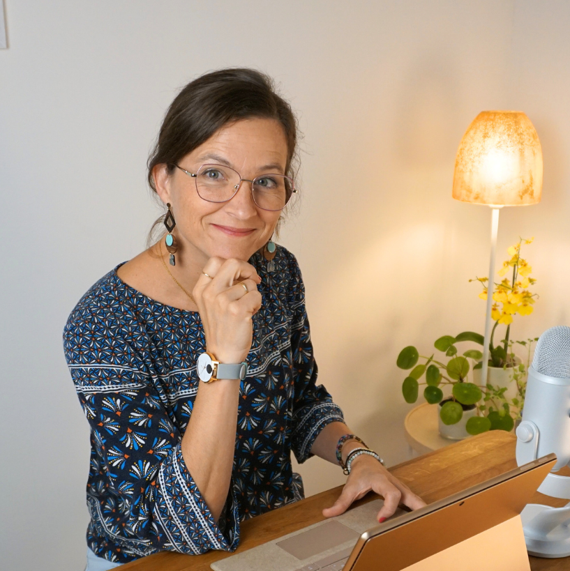 Frau mit Brille und blau gemustertem Oberteil lächelt, arbeitet an einem Laptop am Schreibtisch, die Lampe ist an.