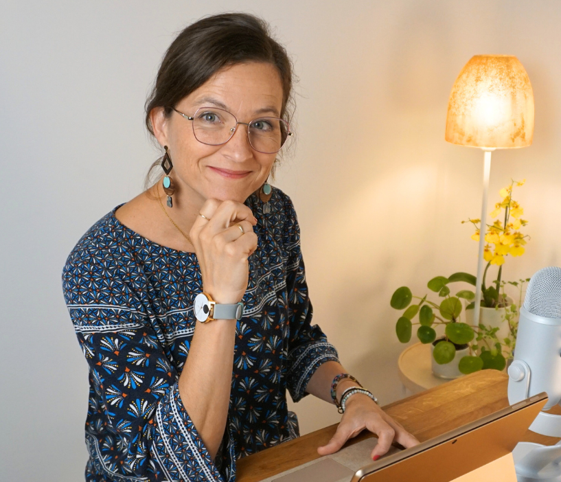 Eine Frau mit Brille sitzt lächelnd mit der Hand am Kinn an einem Schreibtisch; im Hintergrund sind eine Lampe und Blumen zu sehen.