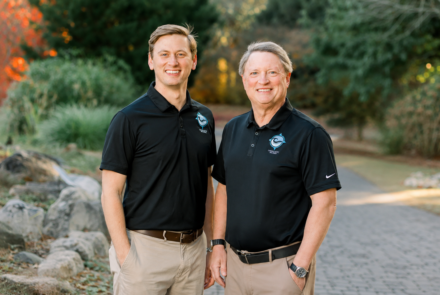 Two men in black polos and khaki pants standing outdoors on a trail, smiling at the camera.