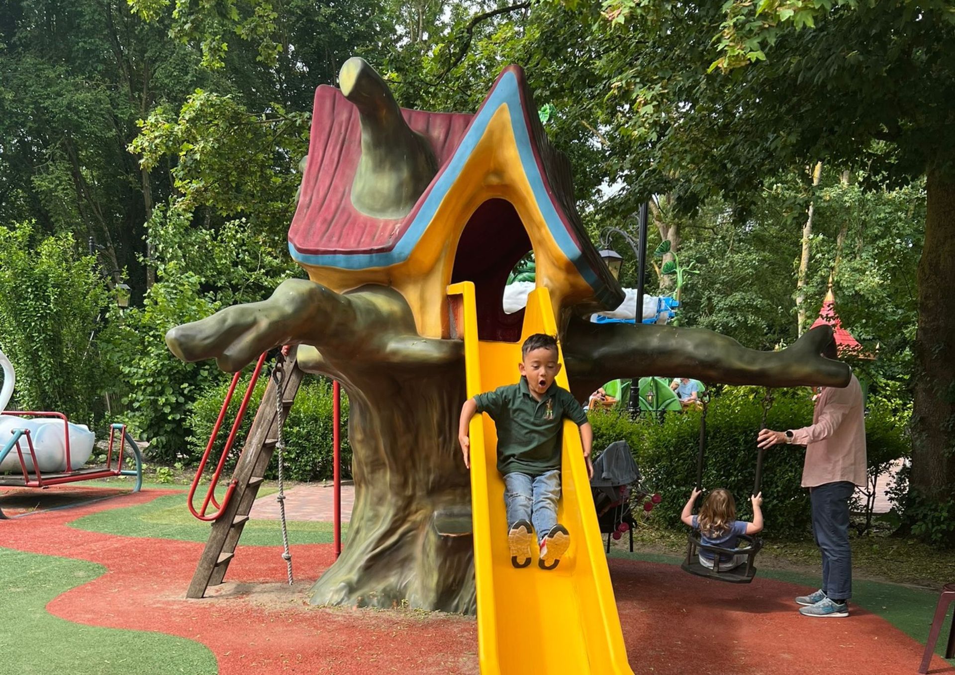 Een kind glijdt van een gele glijbaan op een speelse boomhutconstructie in een park, een ander kind schommelt.